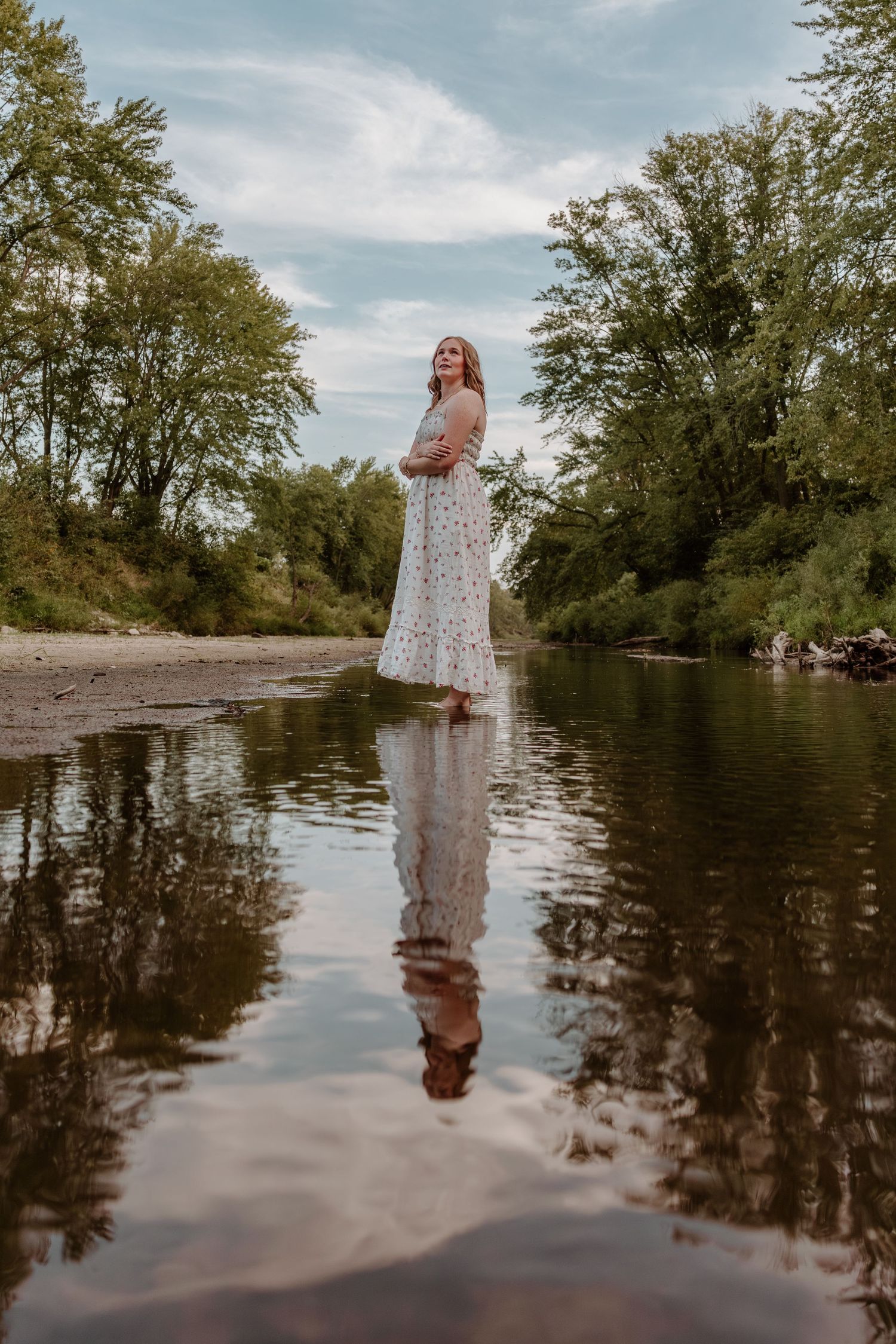 A person in a floral dress stands in shallow water creating mirror-like reflections among trees at sunset.