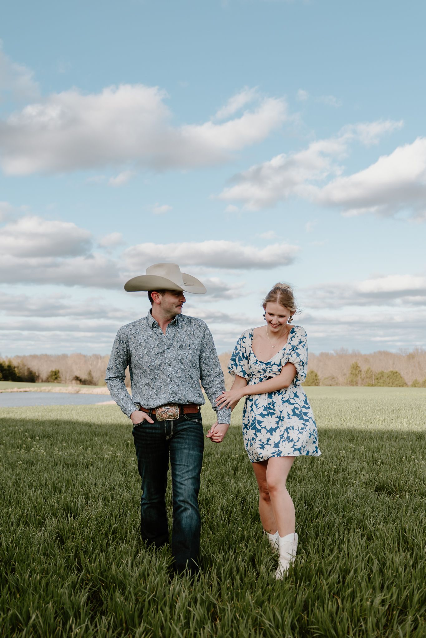 A couple walks hand-in-hand through a green field, man in cowboy hat and floral shirt, woman in floral dress and white boots.