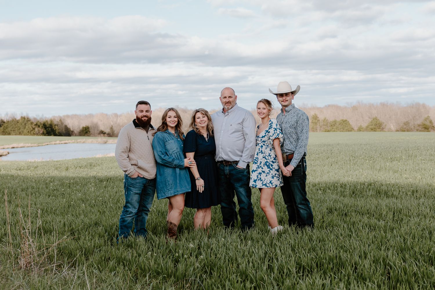 Six people smiling together in a lush green field with mountains and a river in the background.