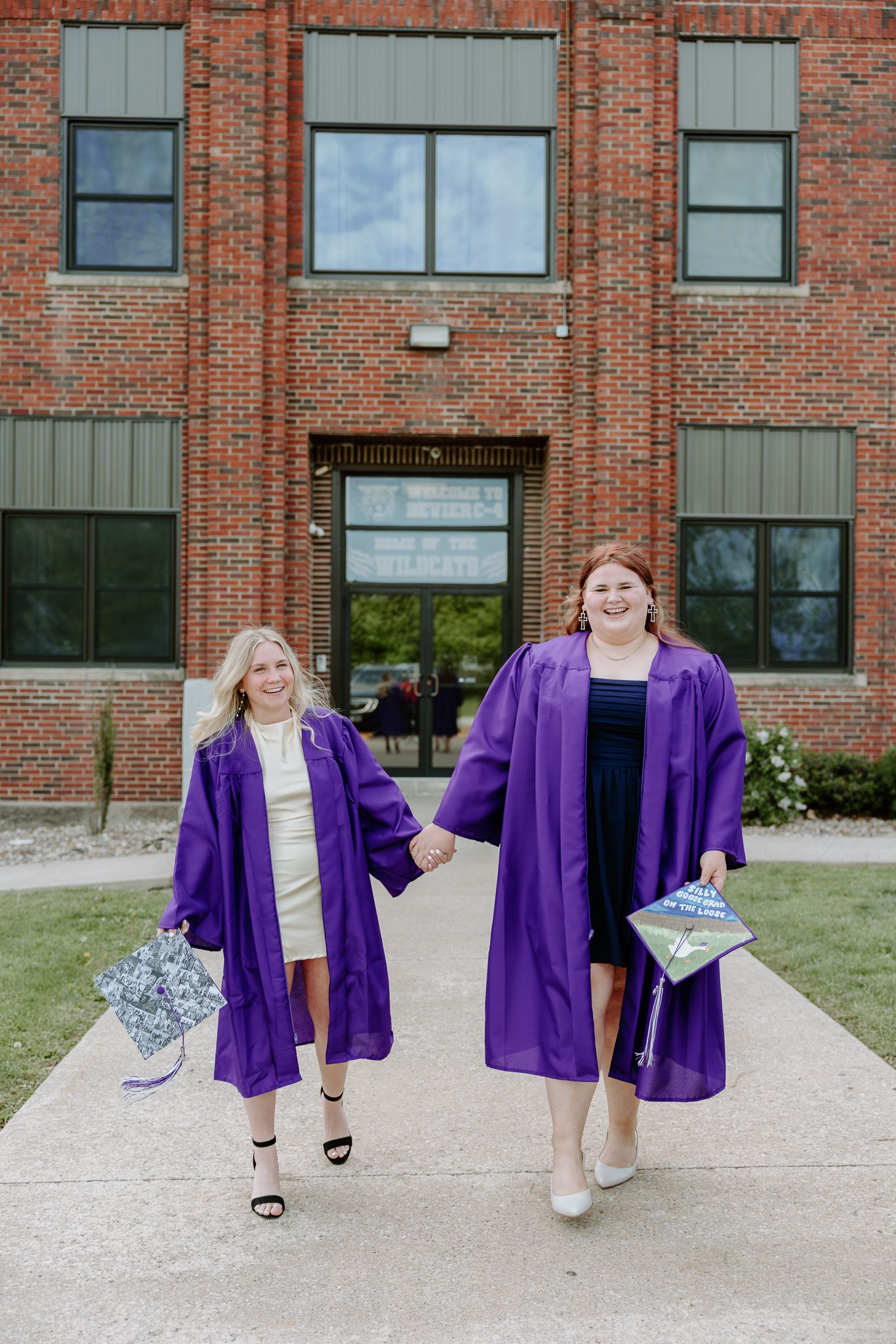 Two women in purple graduation gowns and caps hold hands walking in front of a brick building.