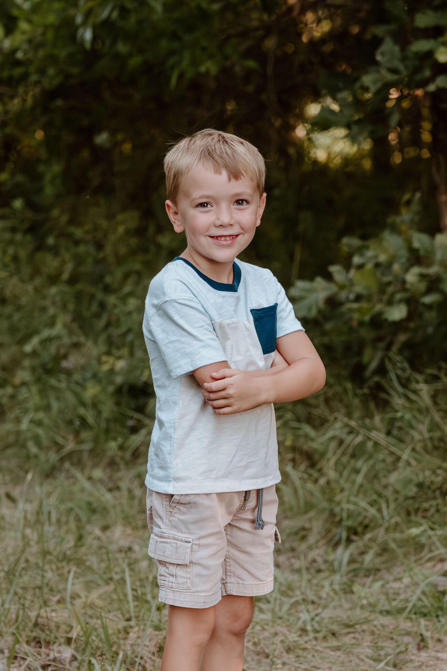 Young child in a light t-shirt and khaki shorts stands outdoors with arms crossed against a natural green background.