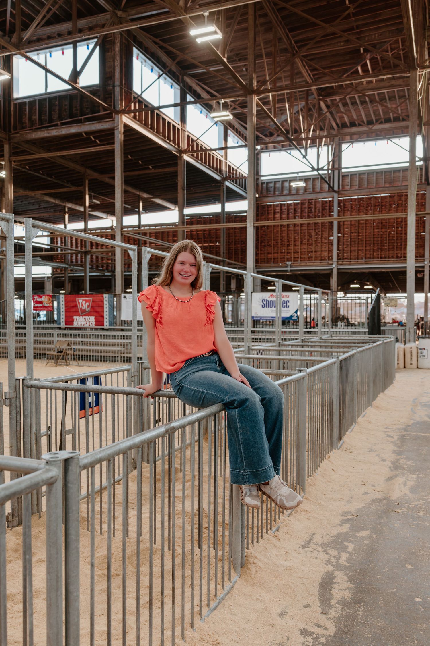 A person in a coral shirt and jeans sits on metal railings inside a large barn or livestock facility structure.