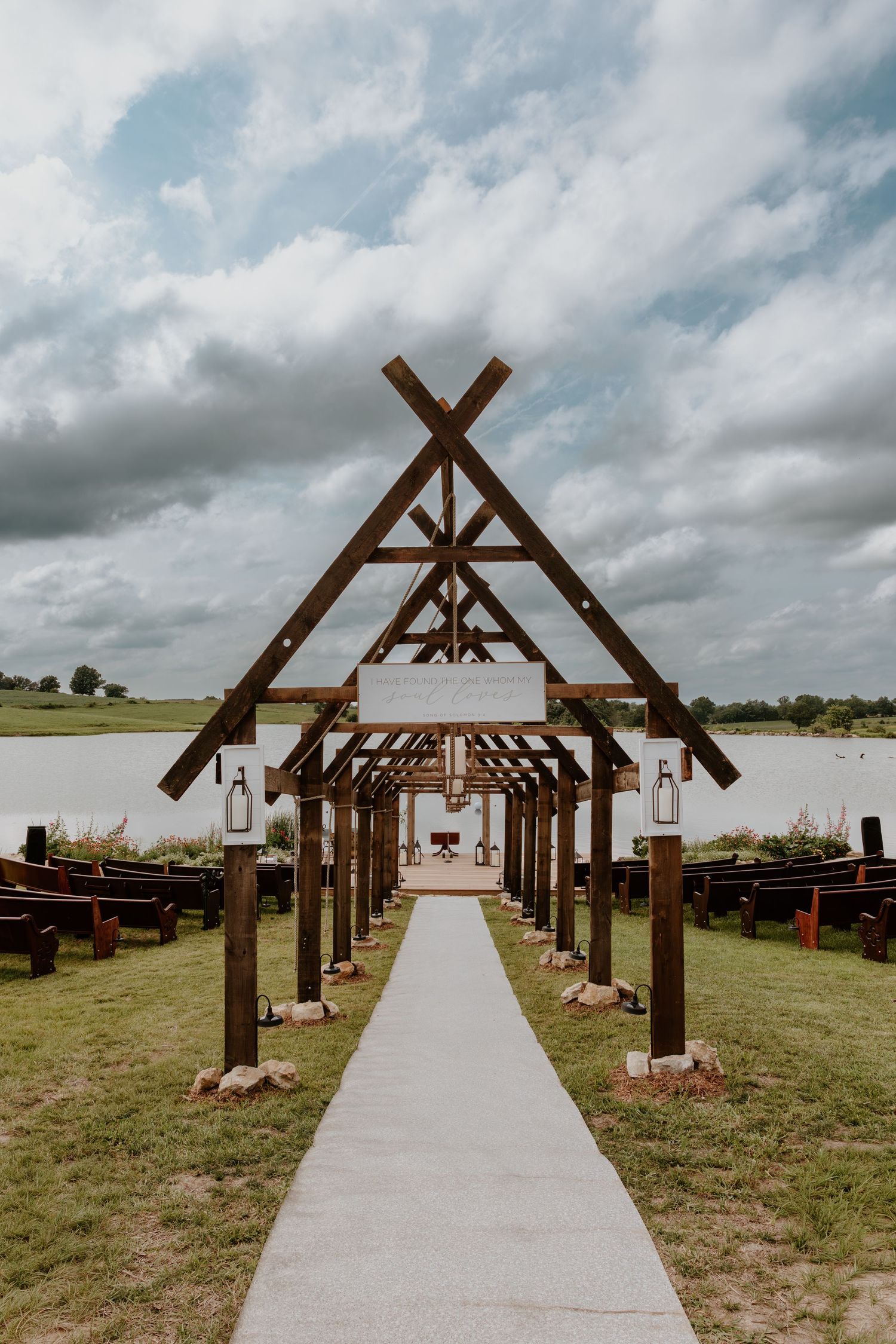 Rustic outdoor wedding ceremony aisle with wooden A-frame arch, lanterns, and pew seating beside a serene lake.