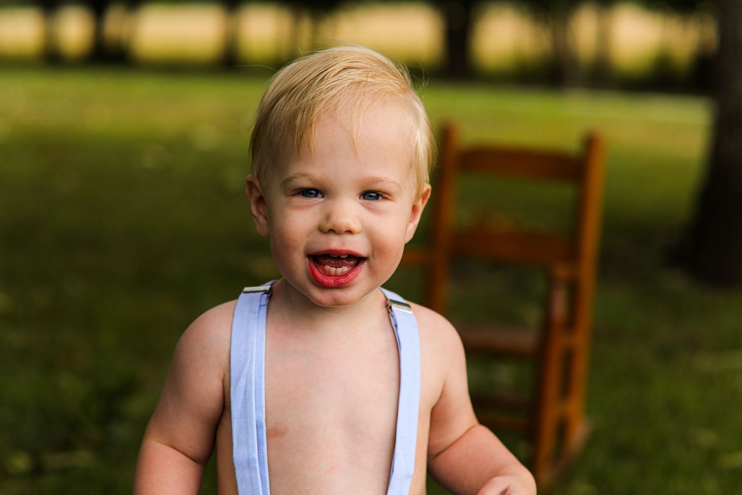 Joyful toddler wearing light blue suspenders smiles in outdoor garden setting.