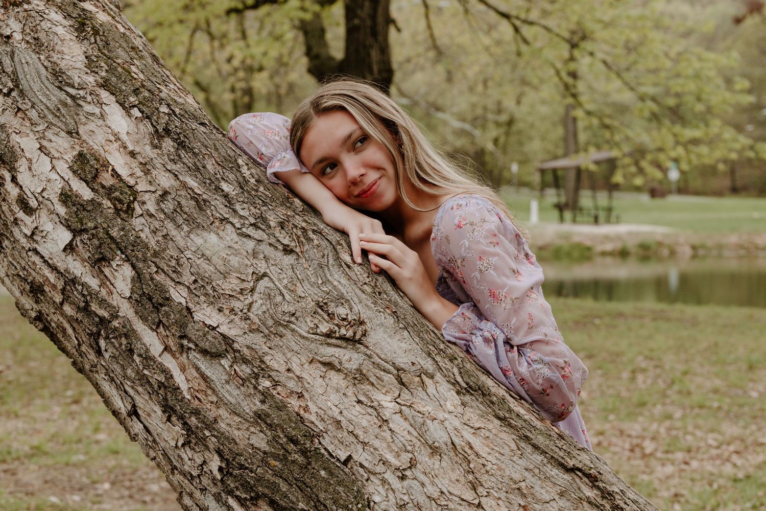 A dreamy portrait of a person in a lace dress leaning against a large tree trunk near a pond on a spring day.