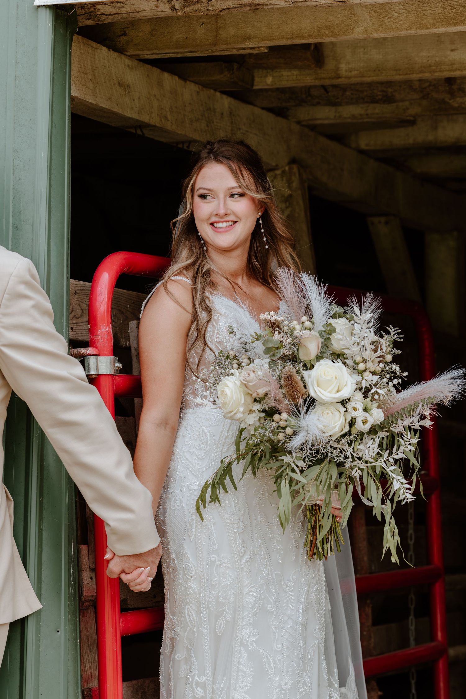 Radiant bride in lace wedding gown holds large bohemian bouquet with white roses and pampas grass in rustic barn setting.