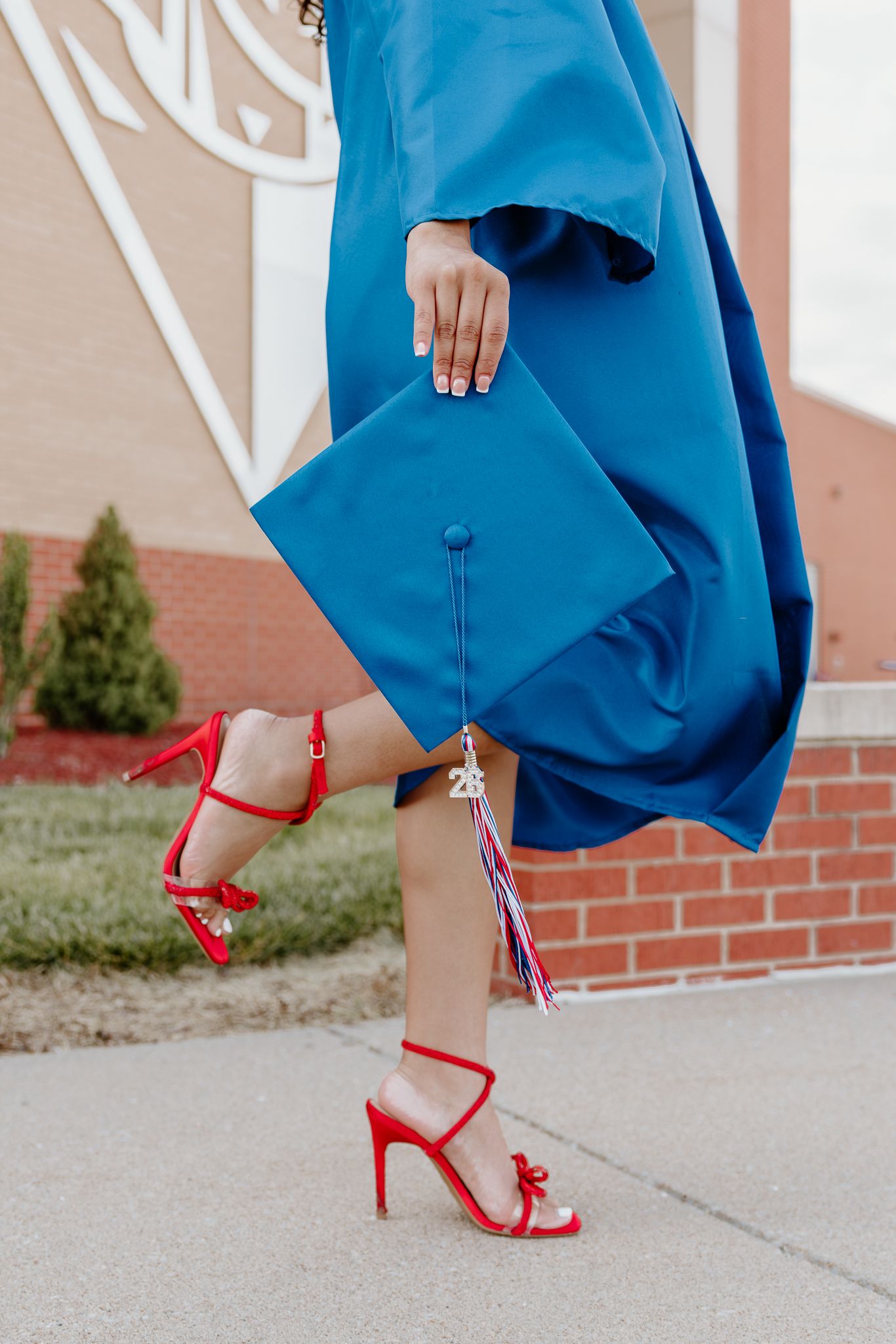 Graduate in blue cap and gown holding mortarboard, wearing red strappy heels on brick school campus.