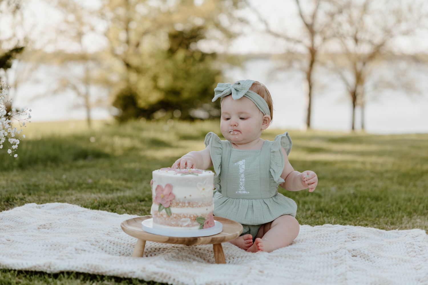 Baby girl in green dress touches first birthday cake outdoors on blanket during golden hour cake smash photo session.