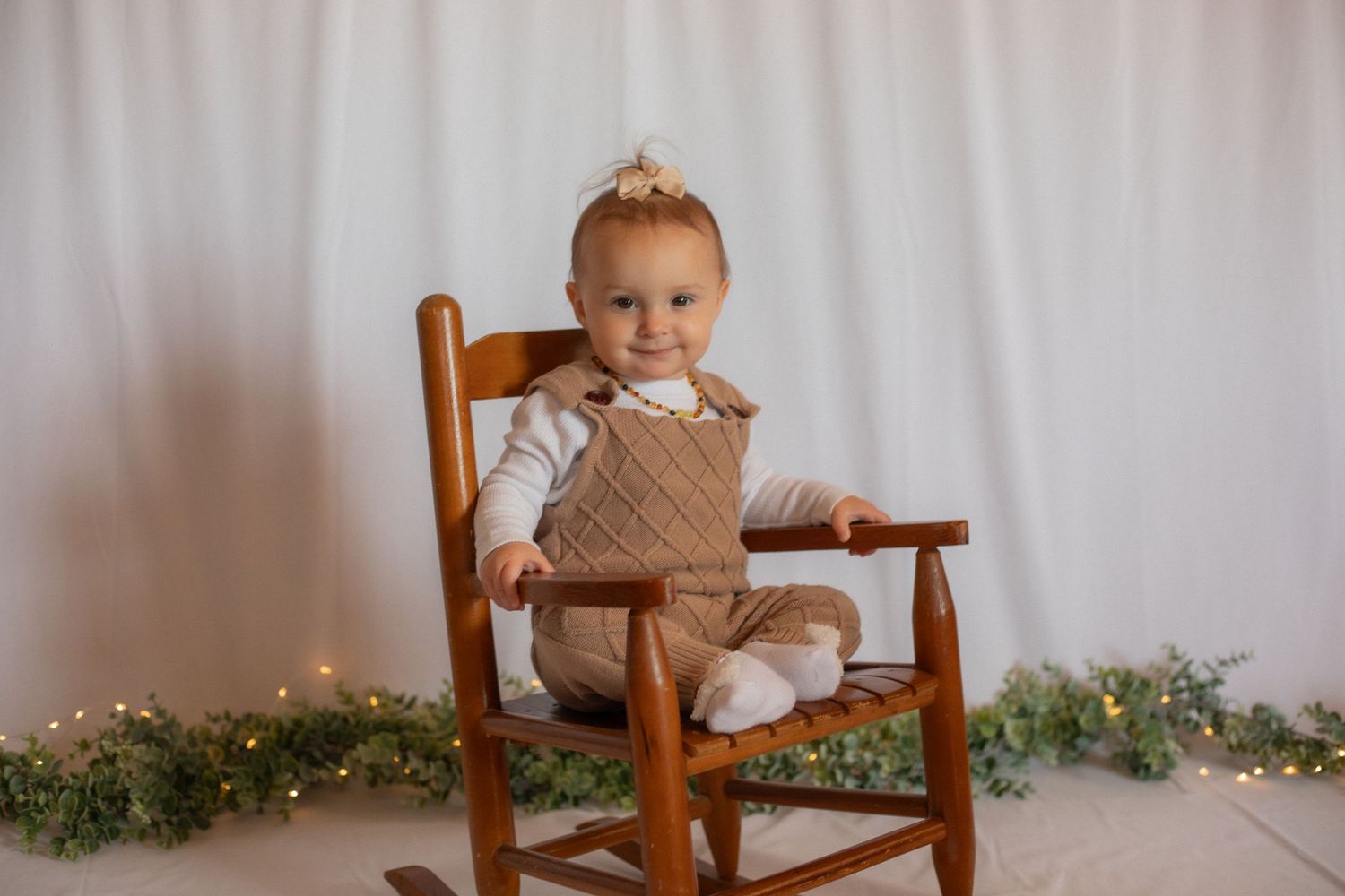 A toddler wearing a brown knit outfit sits in a wooden rocking chair against a white backdrop with twinkle lights.