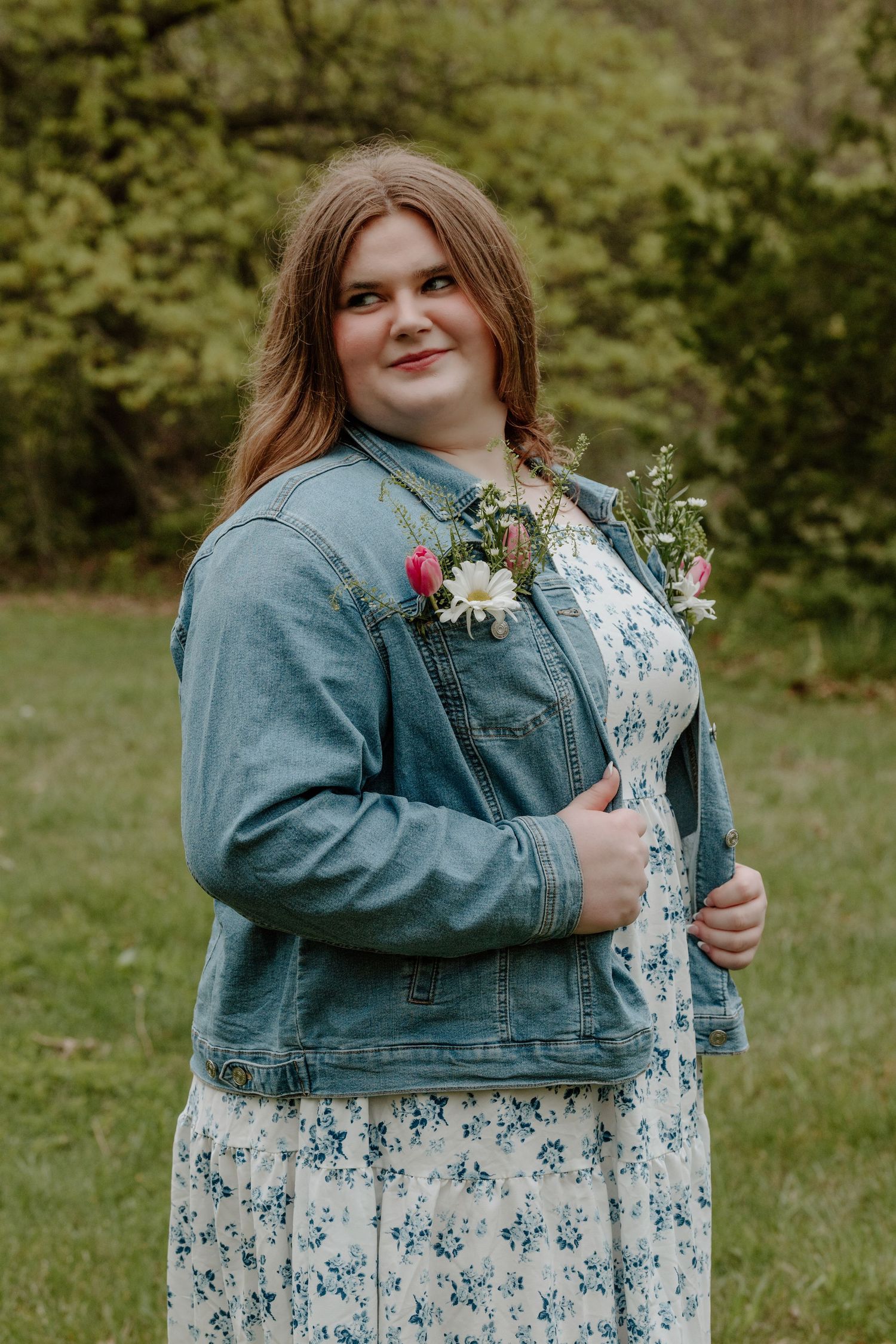 Person in denim jacket and floral dress poses outdoors with spring flowers against green natural background.