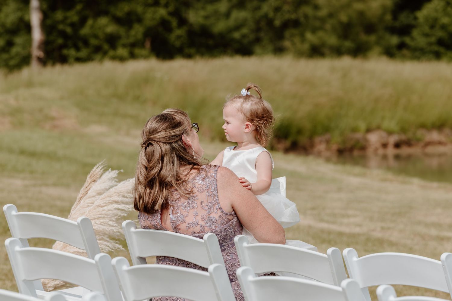 Woman holds young flower girl in white dress while seated at outdoor wedding ceremony chairs.