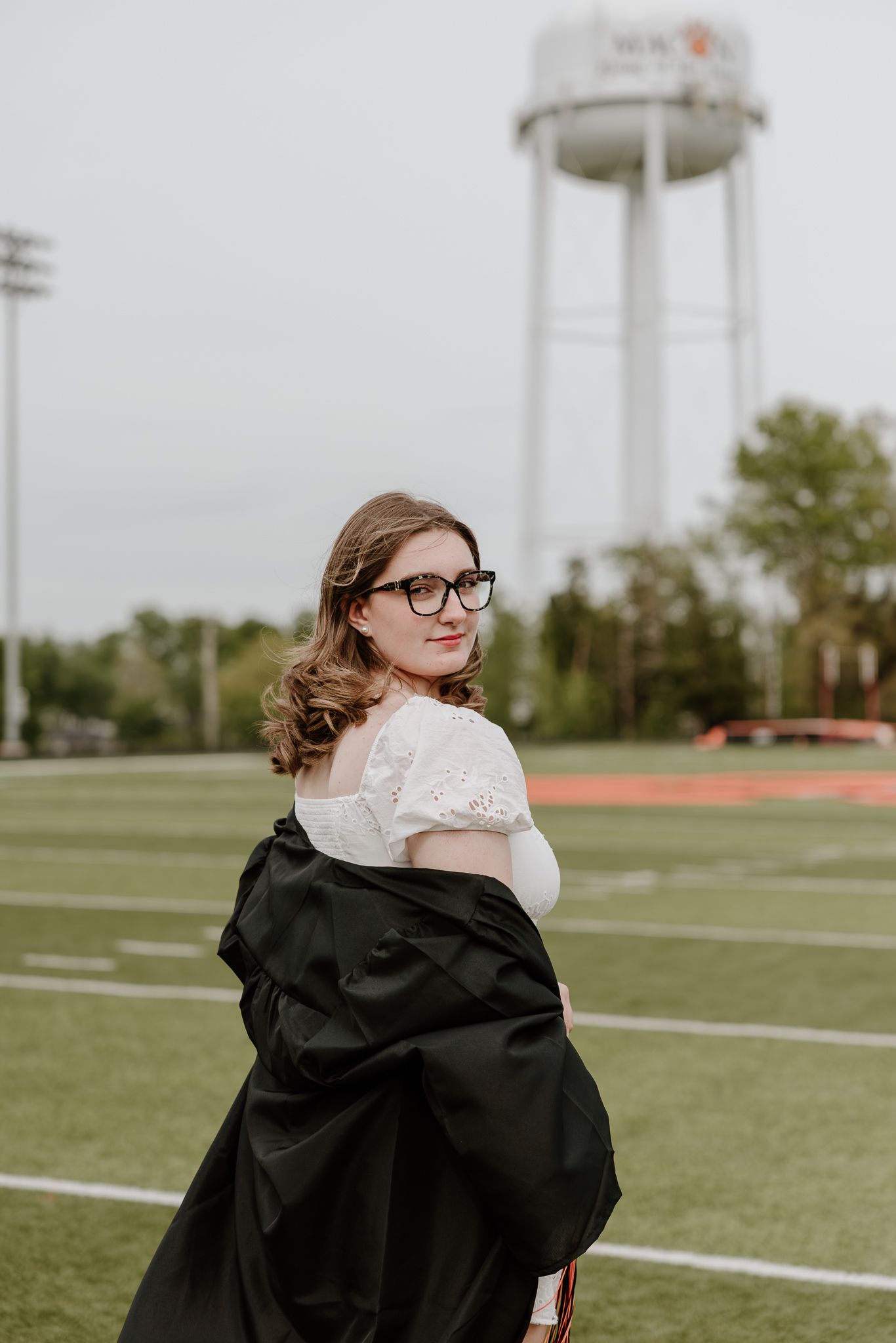 Young woman in graduation gown and glasses poses on athletic field with water tower in background.