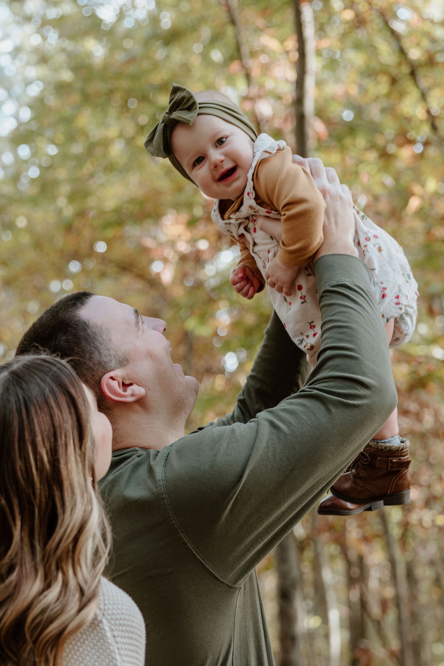 A parent lifts and plays with a small child wearing floral clothing during an autumn photoshoot in a wooded setting.