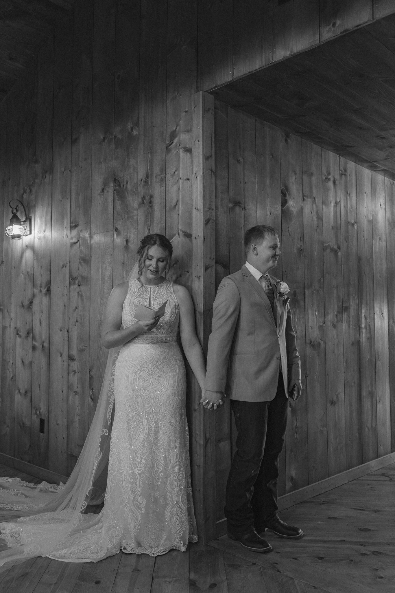 Black and white photo of bride and groom holding hands back-to-back against rustic wood wall.