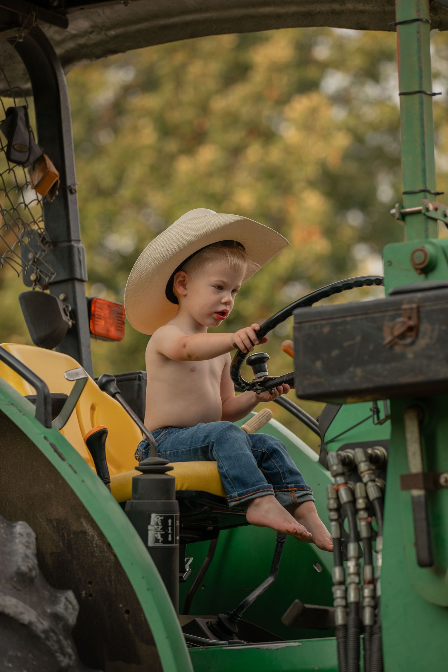 Small child in cowboy hat sits playfully at controls of vintage green tractor.