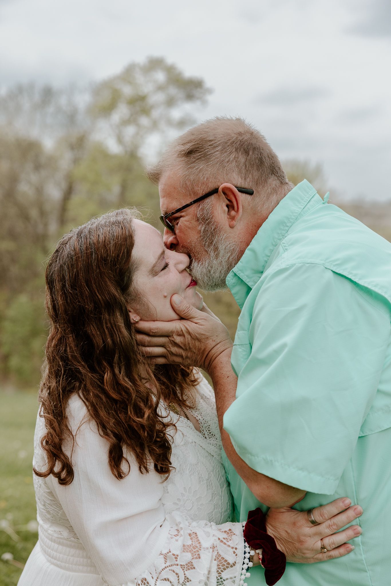 Couple sharing a romantic kiss outdoors, woman in white lace dress and man in mint shirt, surrounded by green trees.