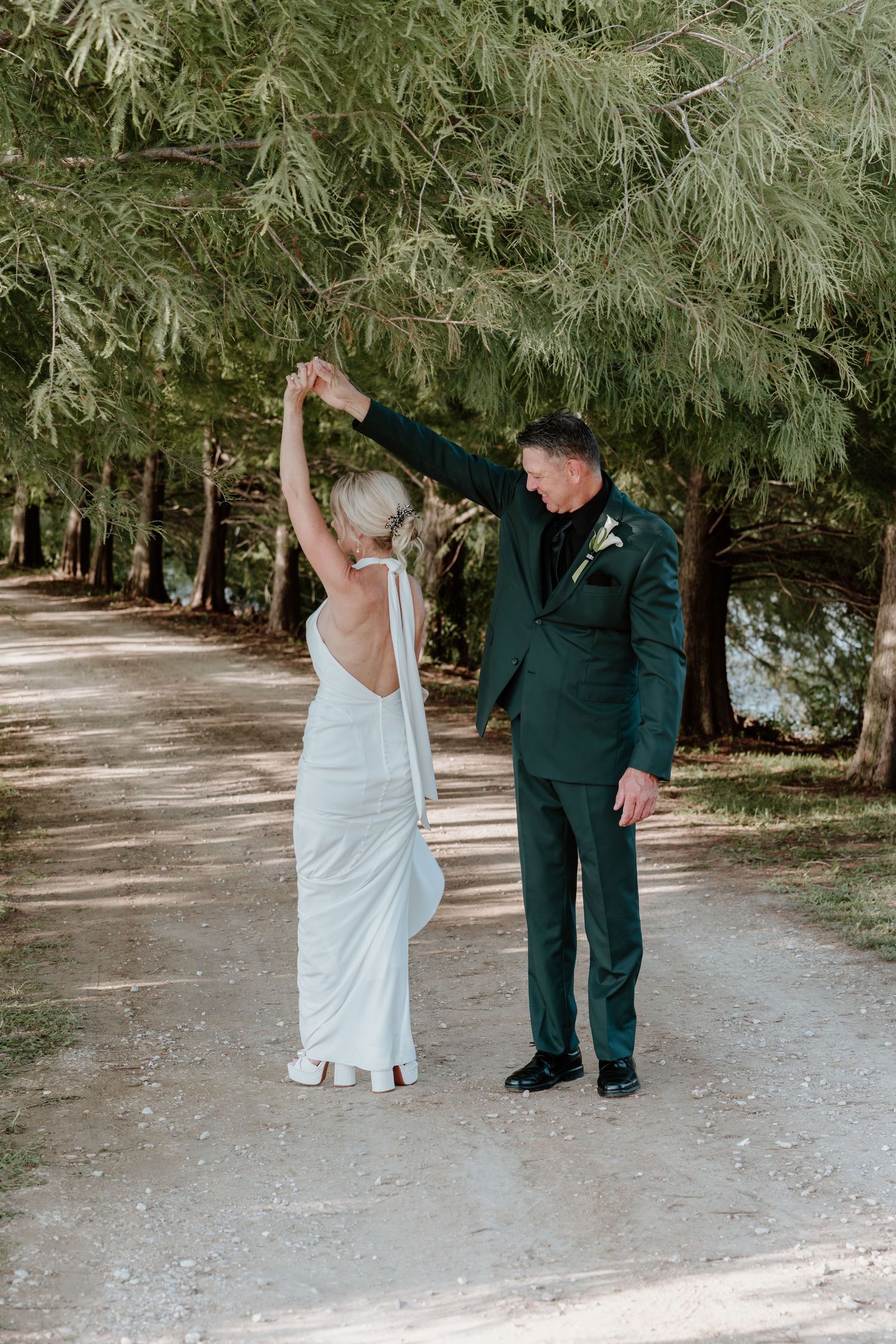 Bride in white gown and groom in green suit dancing on a tree-lined path at an outdoor wedding.