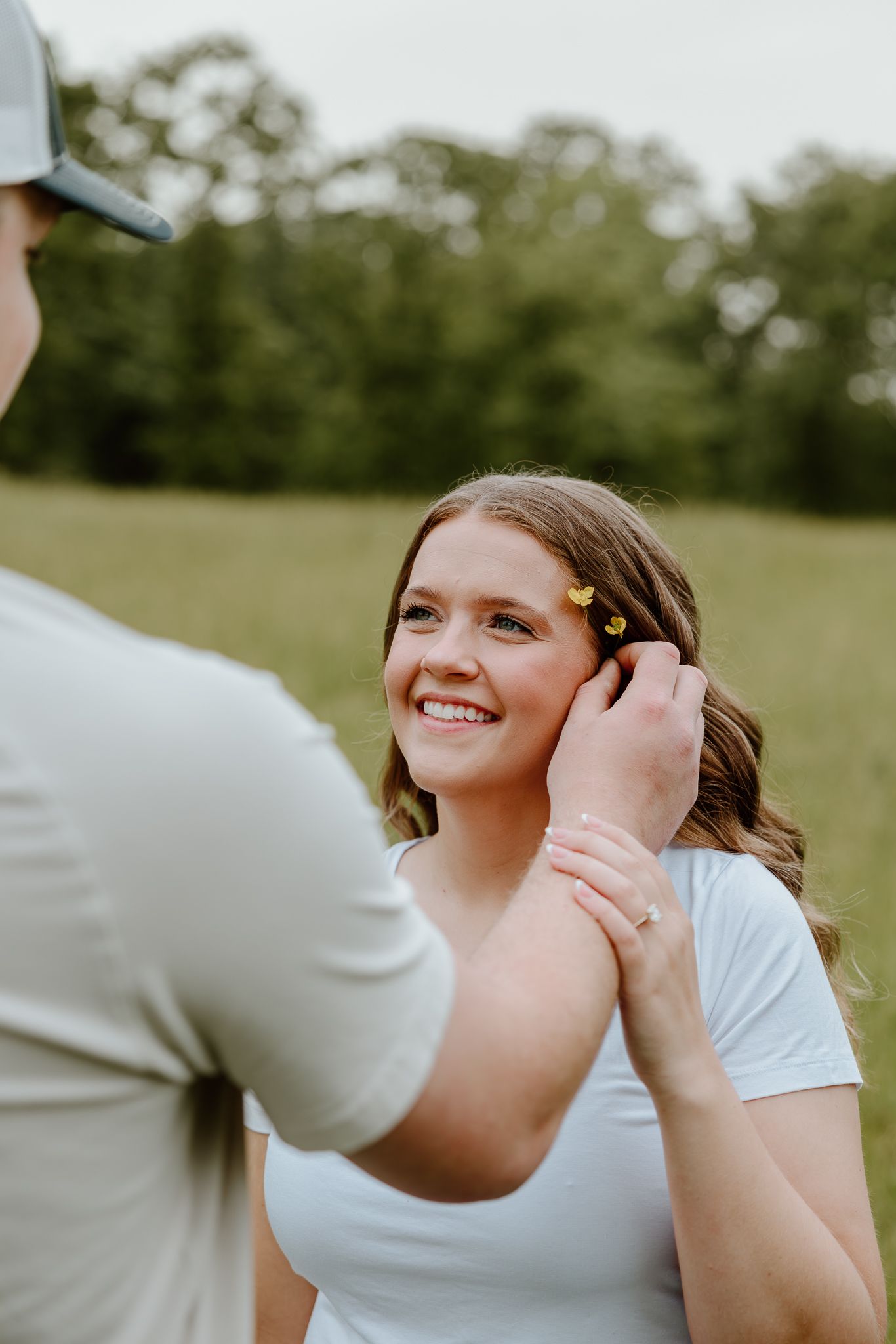 A man places a yellow flower in a smiling woman's hair outdoors in a green field.