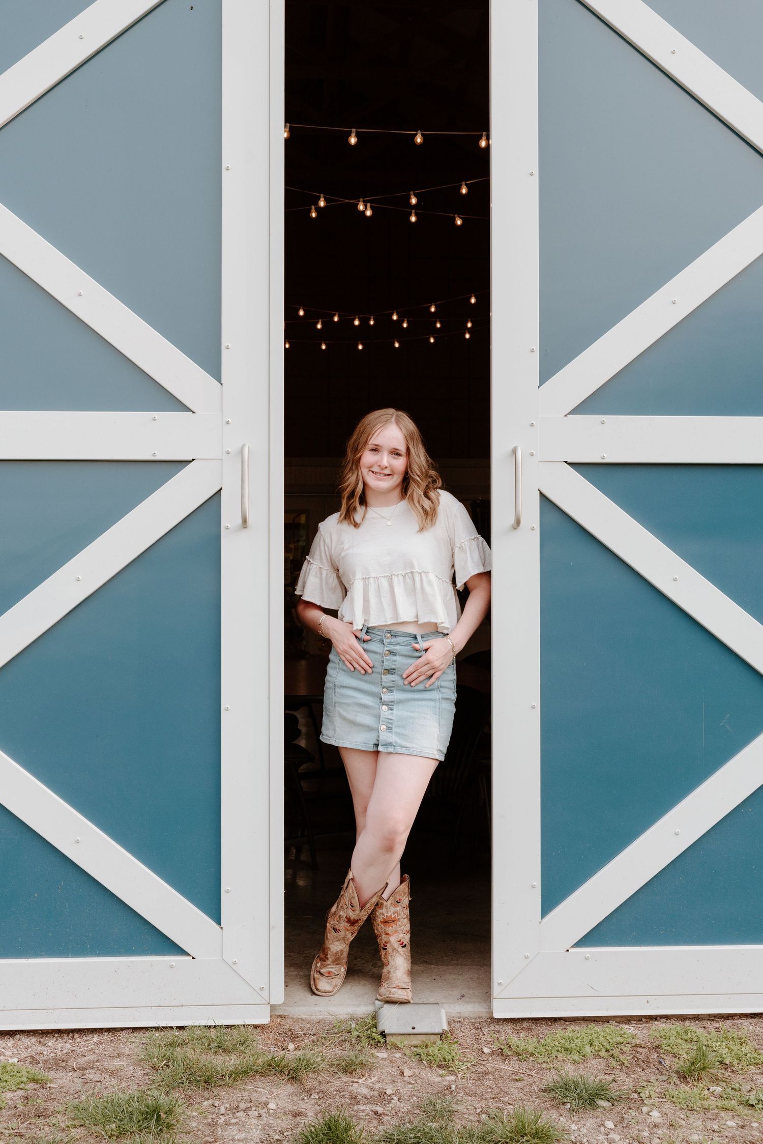 Someone poses in a rustic barn doorway wearing a white top, denim shorts and brown boots against a blue-painted structure.