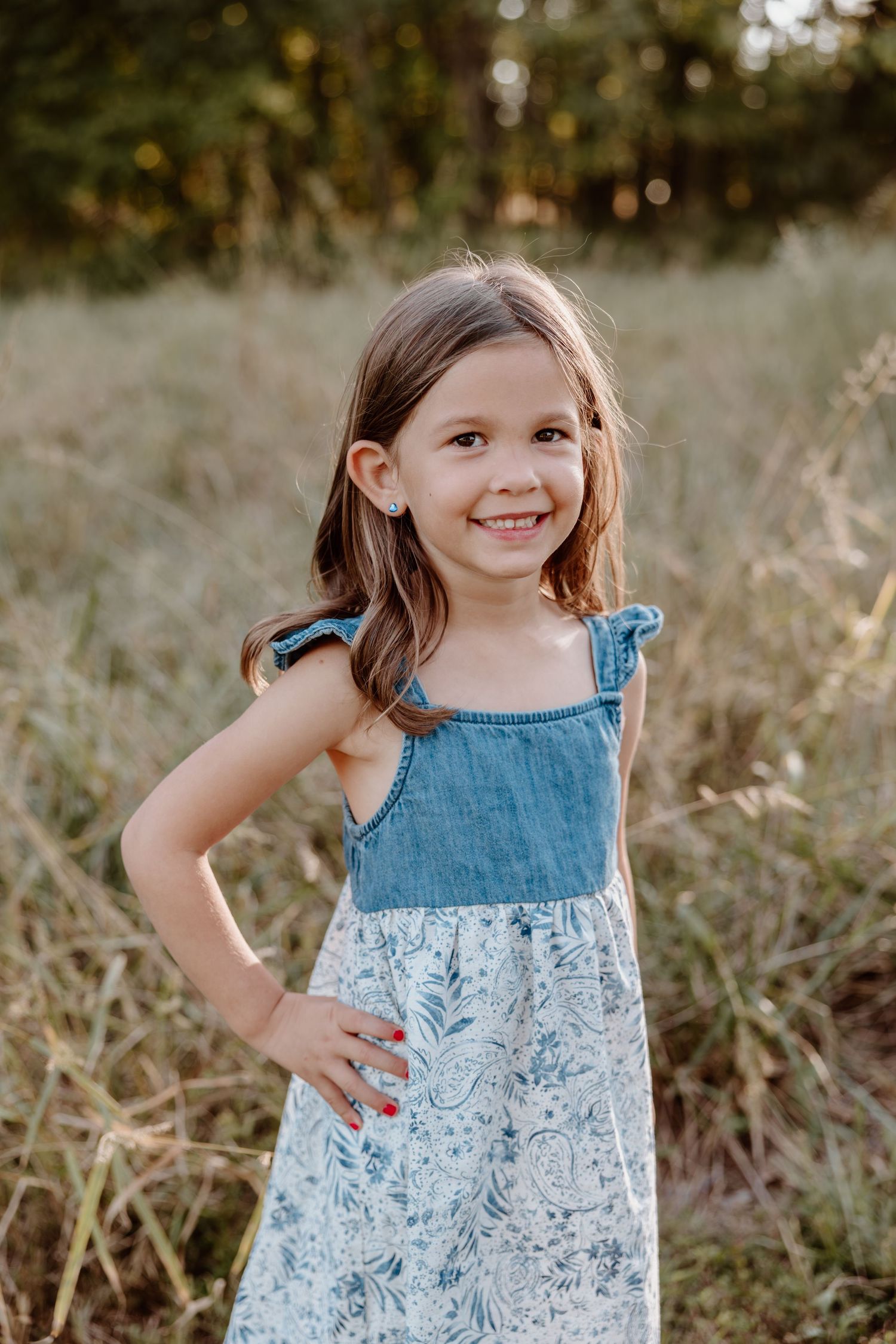 A young child poses in a denim and floral print dress while standing in a grassy field during golden hour.