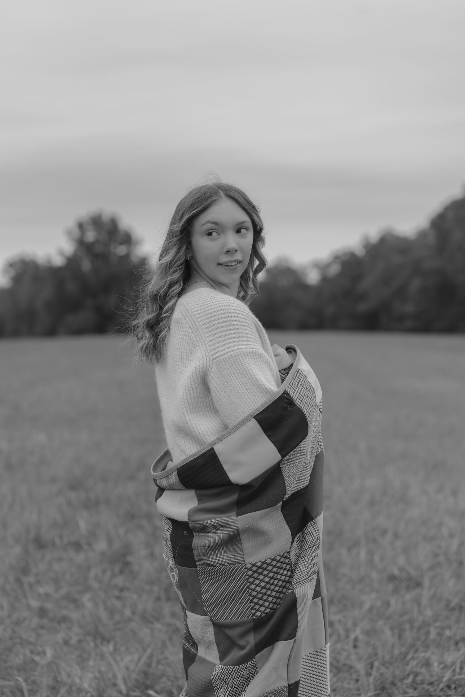 A child wearing a patchwork dress spins playfully in a grassy meadow, captured in black and white photography.