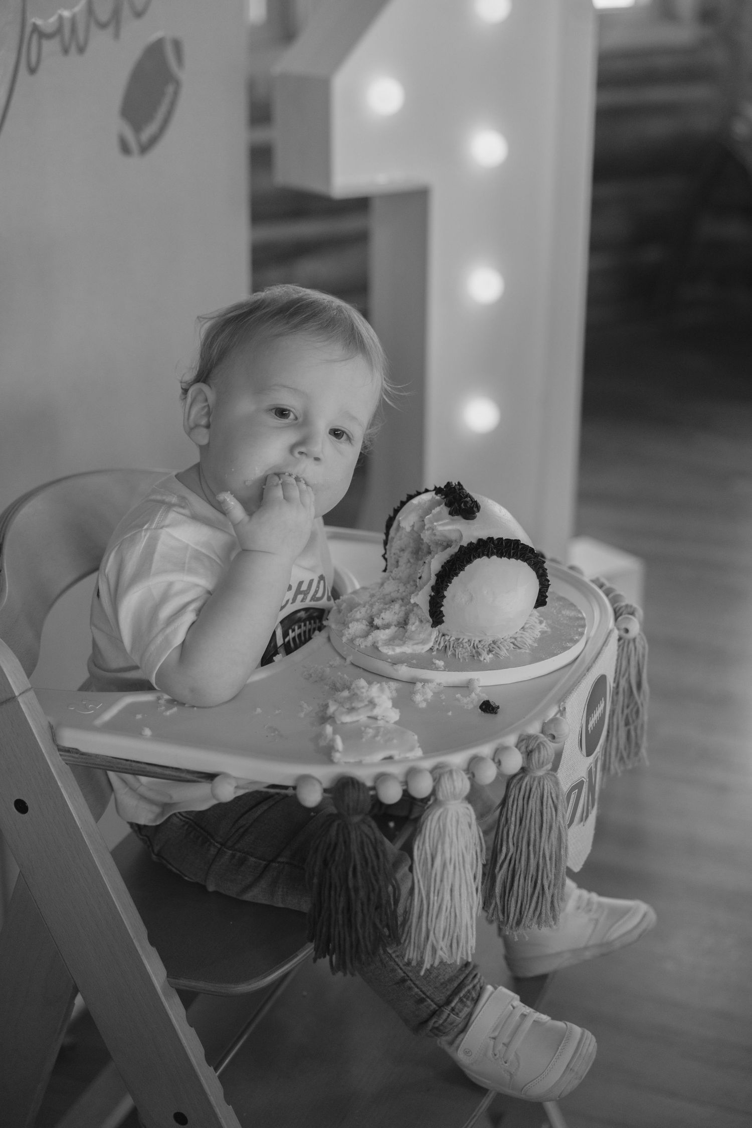 Black and white photo of a toddler in a high chair with a decorated birthday cake, lit by string lights in the background.