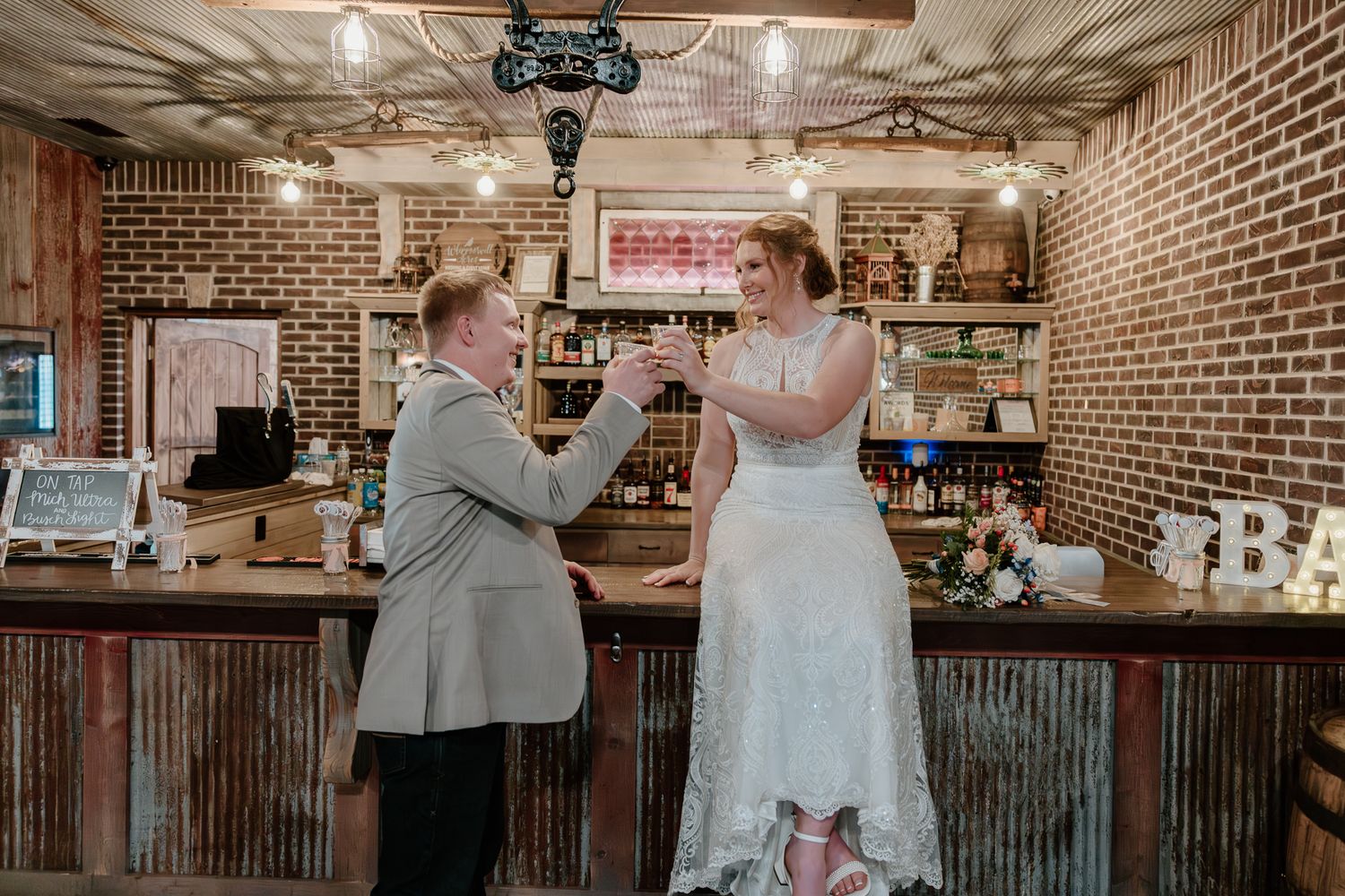 Bride and groom toast drinks at a rustic brick bar decorated with vintage decor on their wedding day.