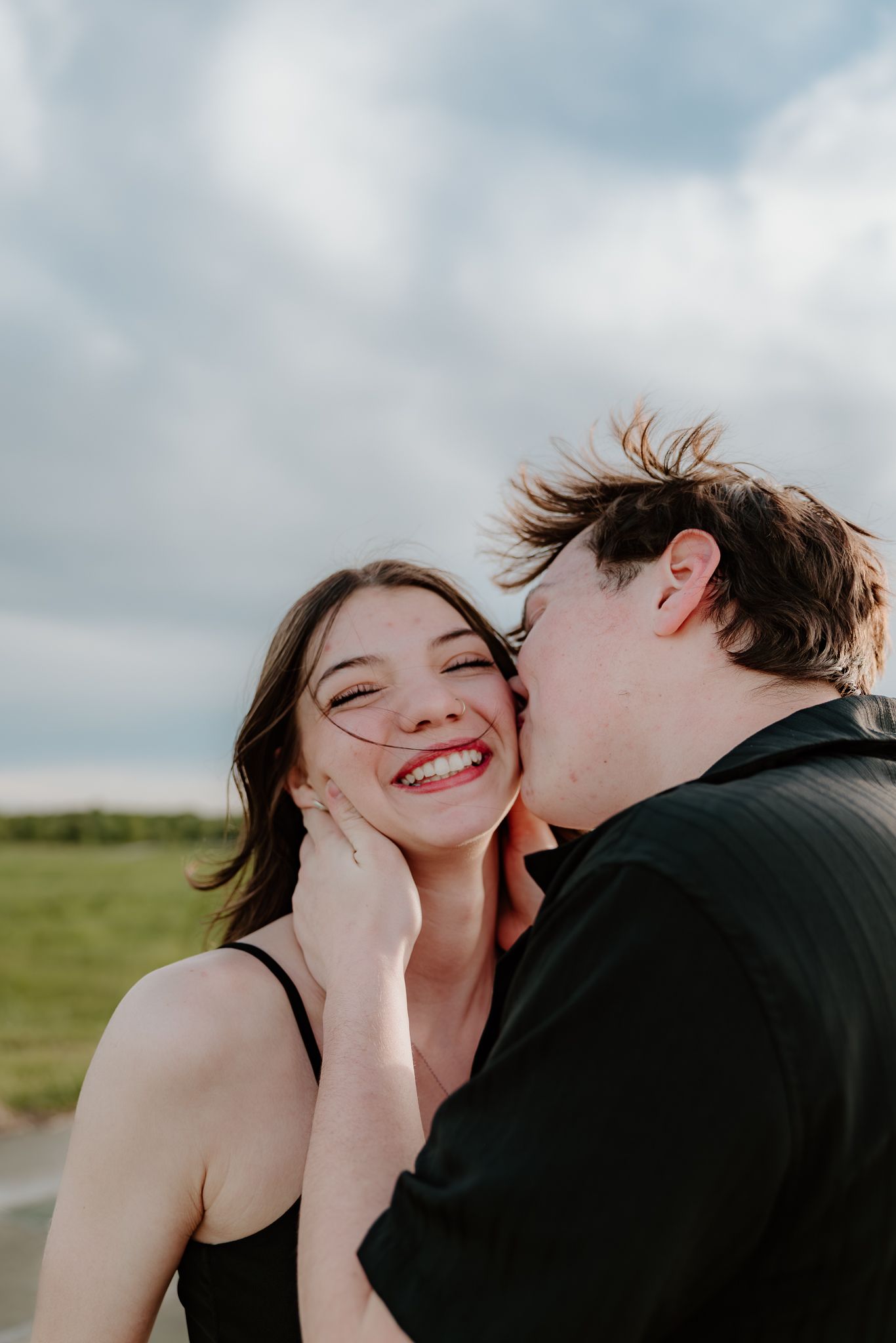 A young couple sharing a romantic moment outdoors, with the man kissing the woman's cheek as she smiles joyfully.