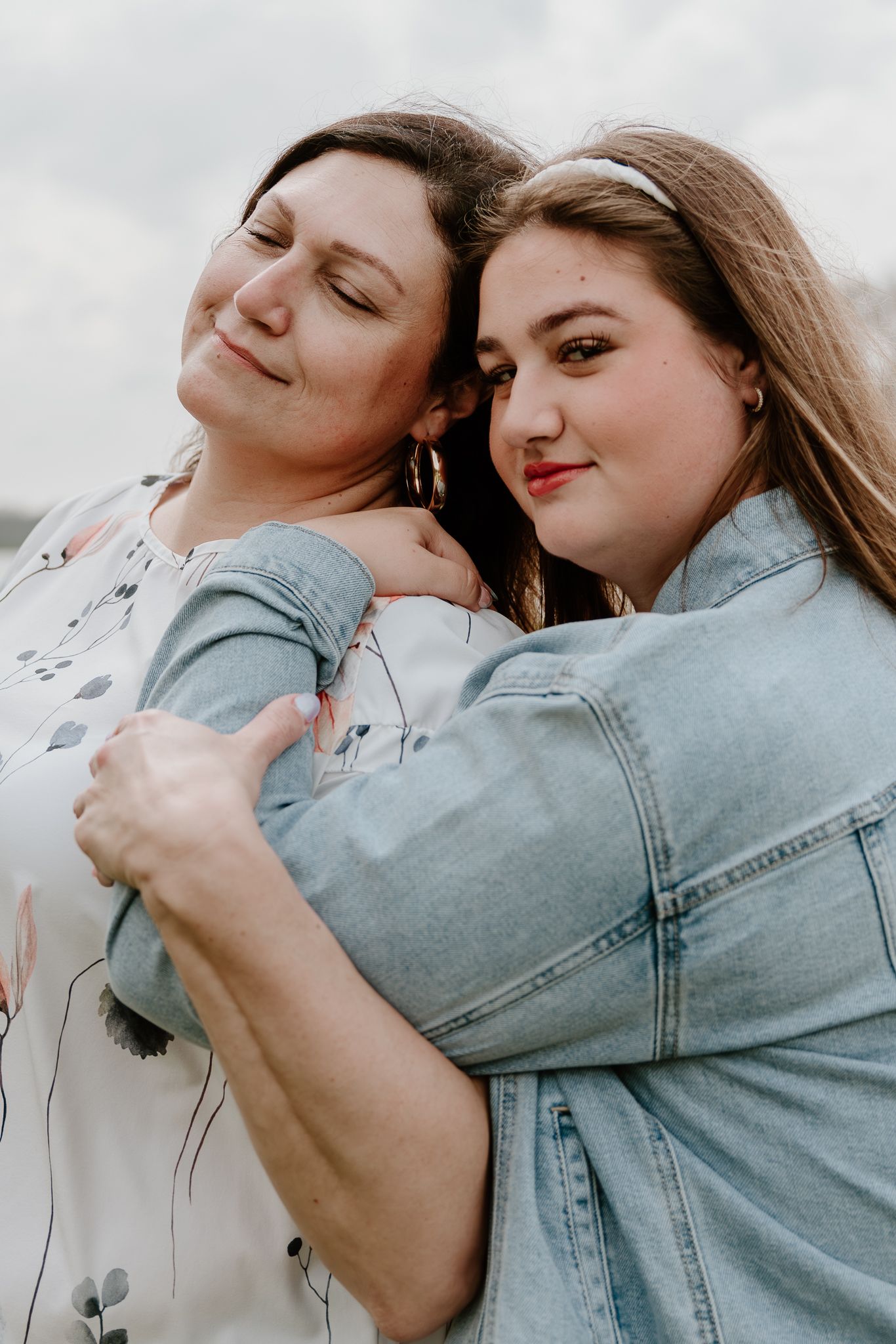 Two women share a warm embrace outdoors, one in a floral top and the other in a denim jacket, smiling contentedly.
