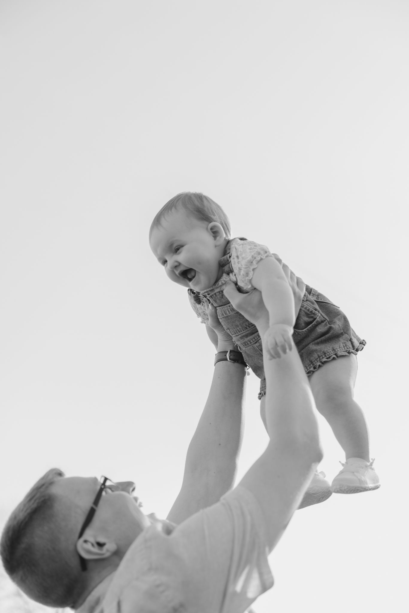 Black and white photo of a laughing baby being lifted overhead by a father outdoors.