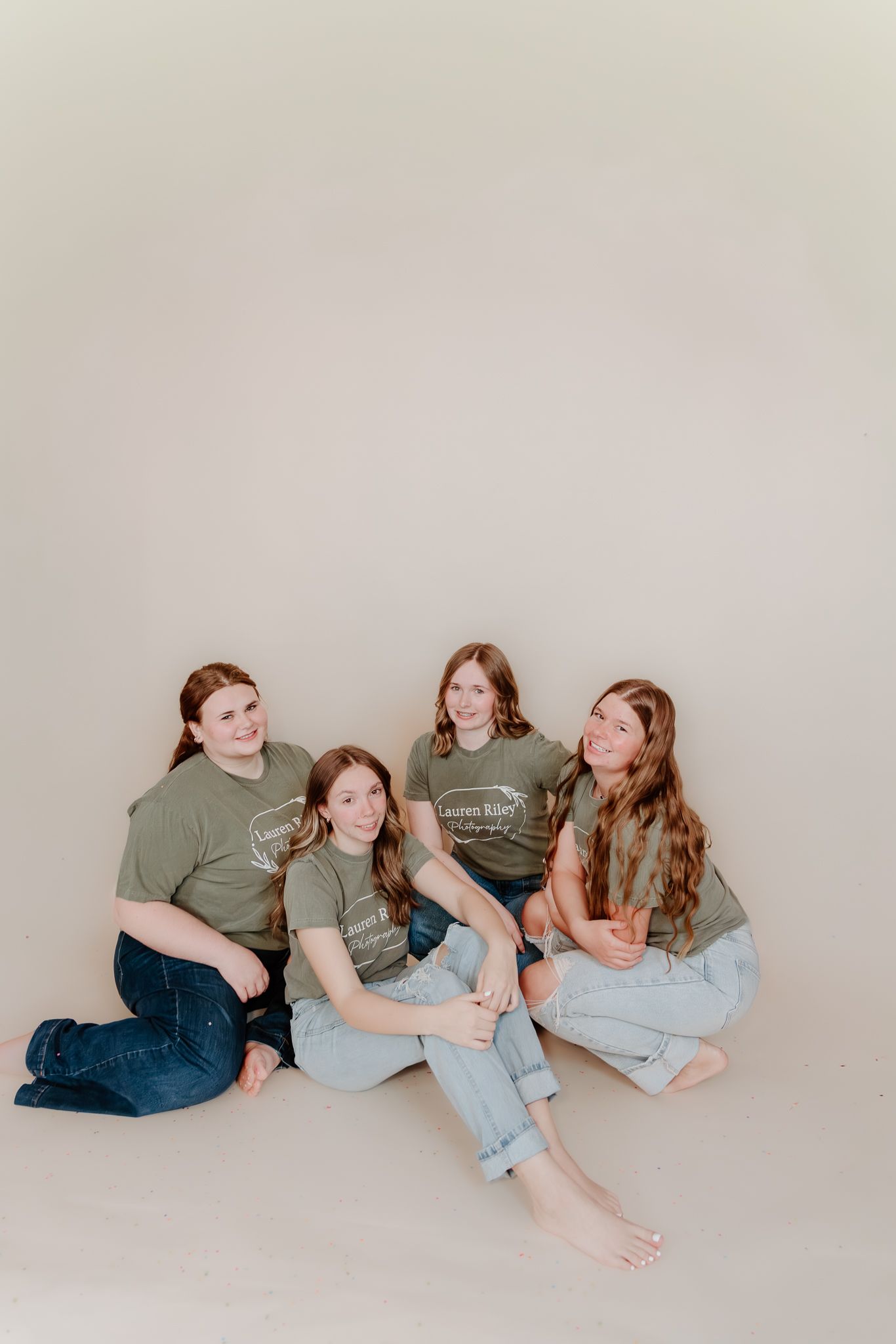 Four girls in matching olive green t-shirts and jeans sitting together on a studio floor, smiling and posing for a group photo.