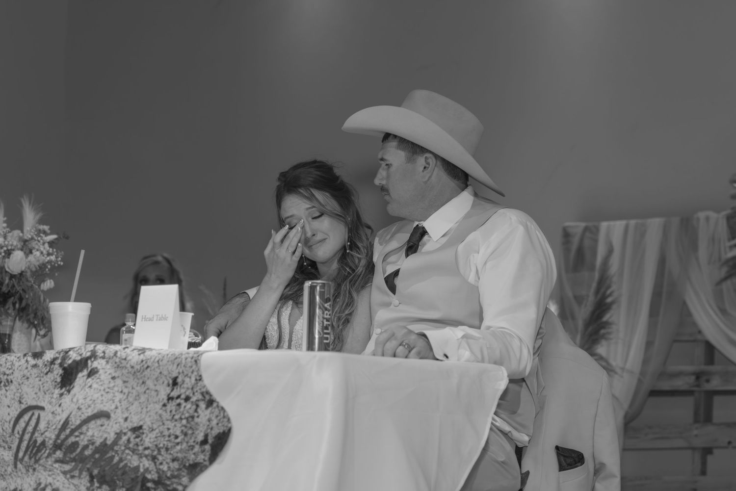 A bride wipes tears at the reception table while seated next to her groom in a white suit and cowboy hat.