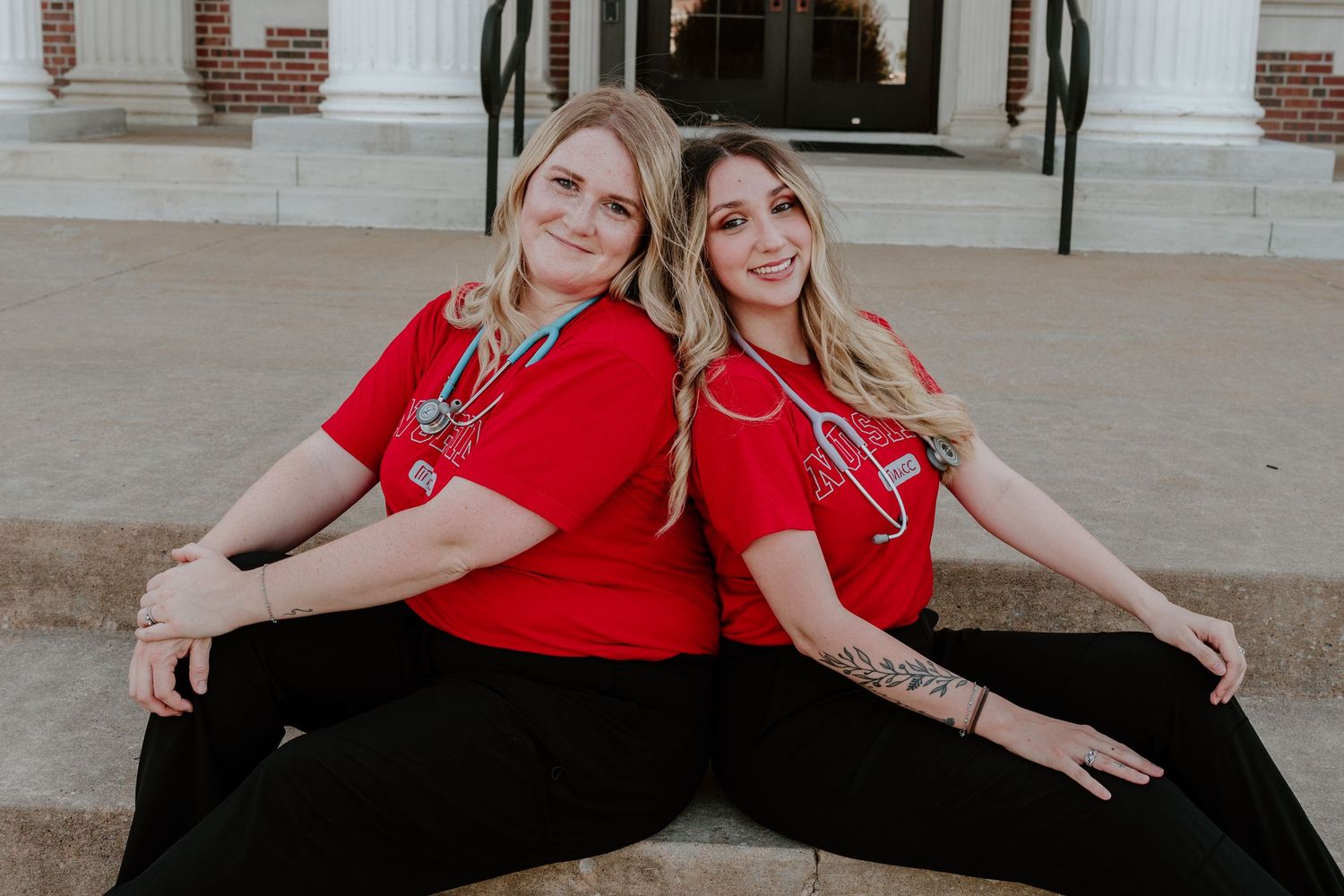 Two women in matching red shirts and black pants sit back-to-back on the ground in front of a brick building.