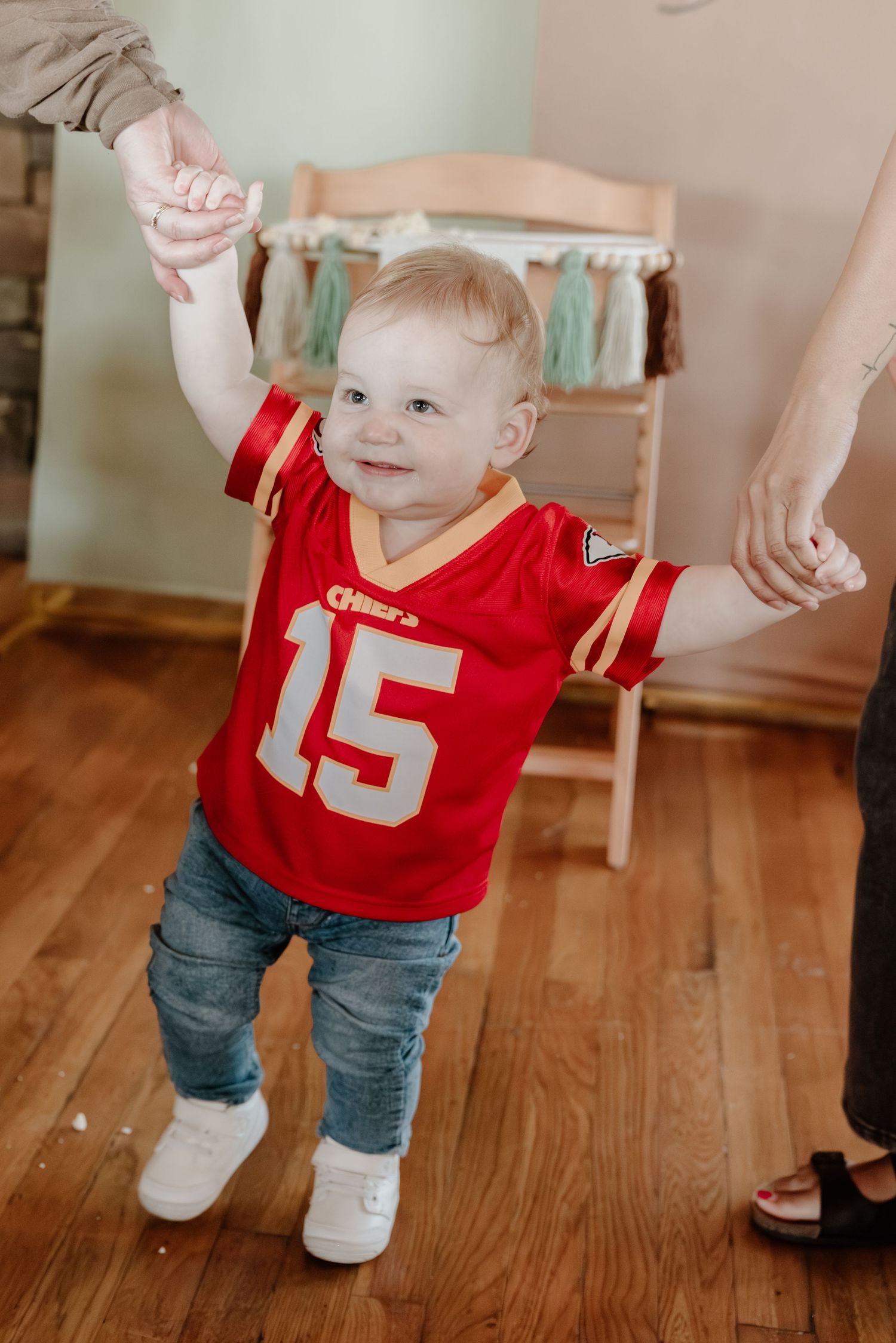 A small child in a red sports jersey and jeans takes first steps while being supported on a hardwood floor.
