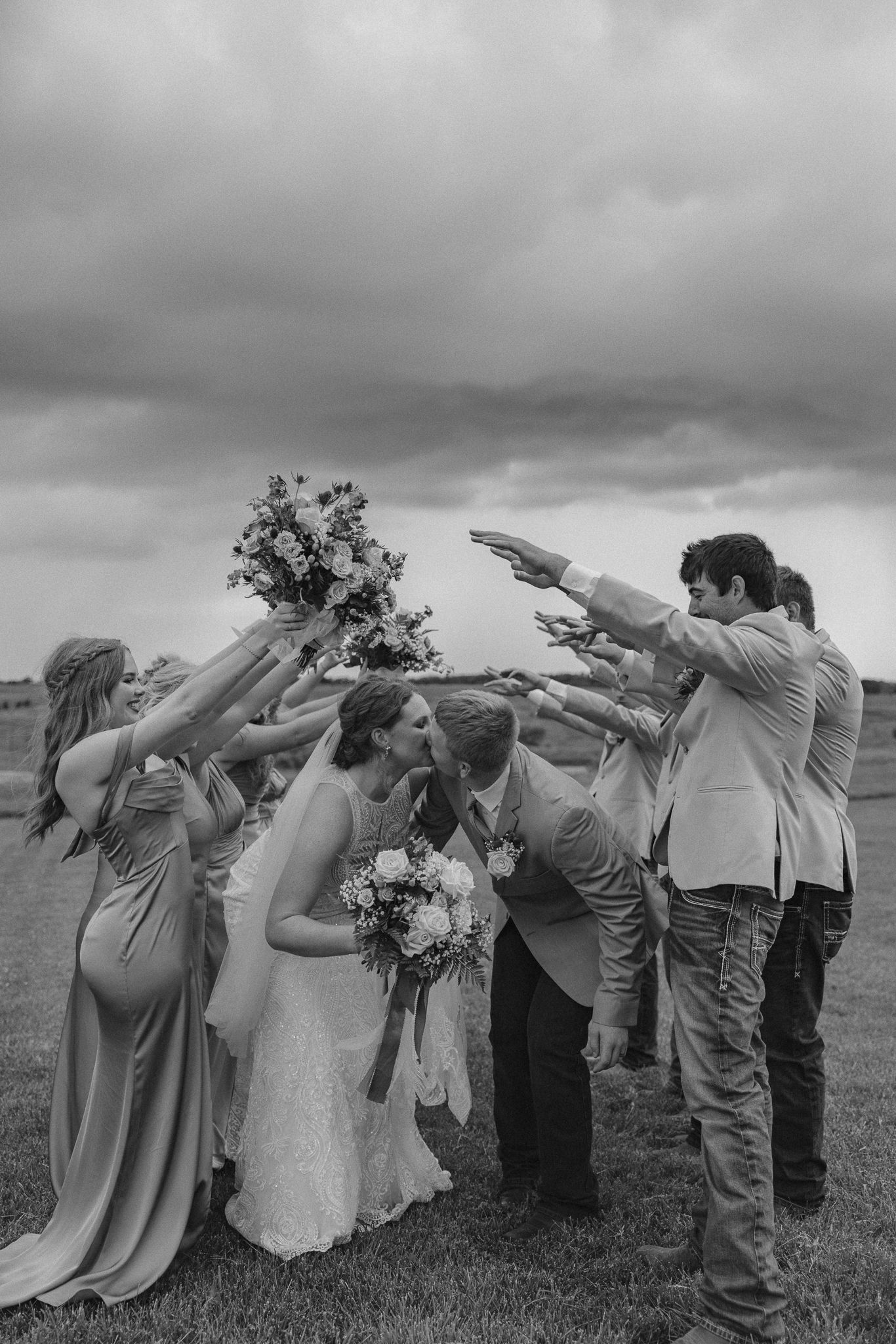 Black and white photo of bride and groom kissing under an arch of raised hands formed by their wedding party outdoors.