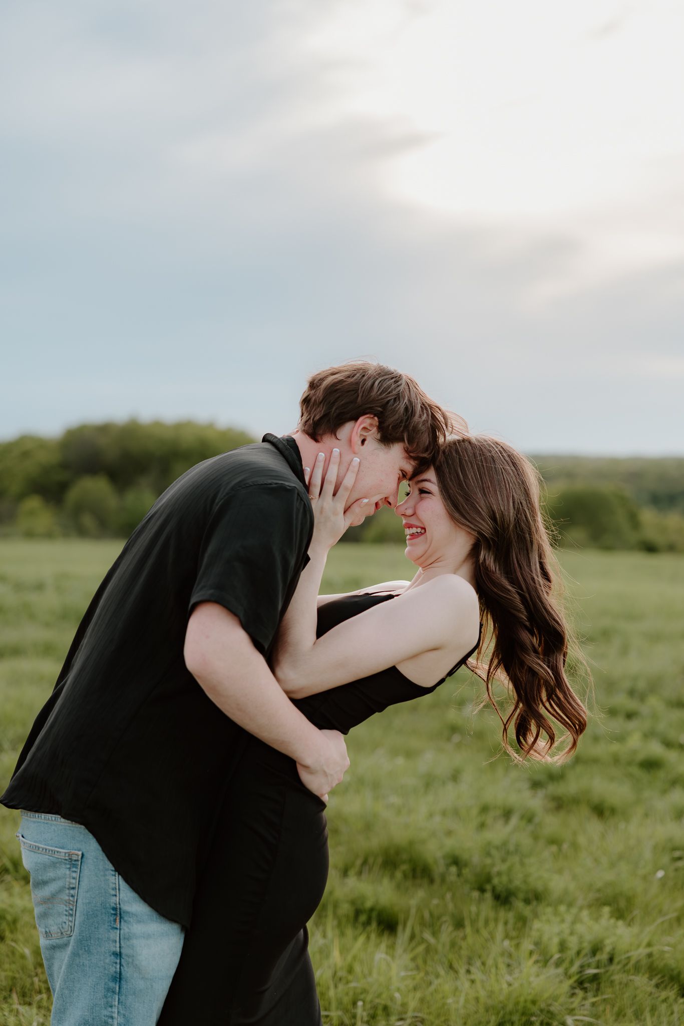 A couple in black outfits share a romantic moment in a lush green field, smiling as they lean in close to kiss.