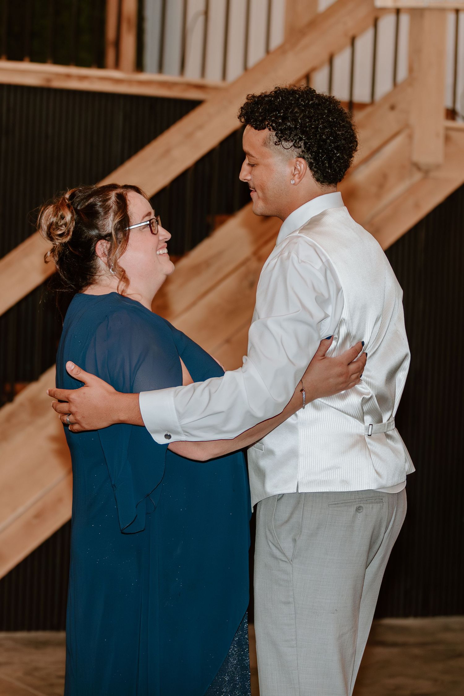 Groom in white shirt dancing joyfully with woman in teal dress near wooden staircase at wedding reception.