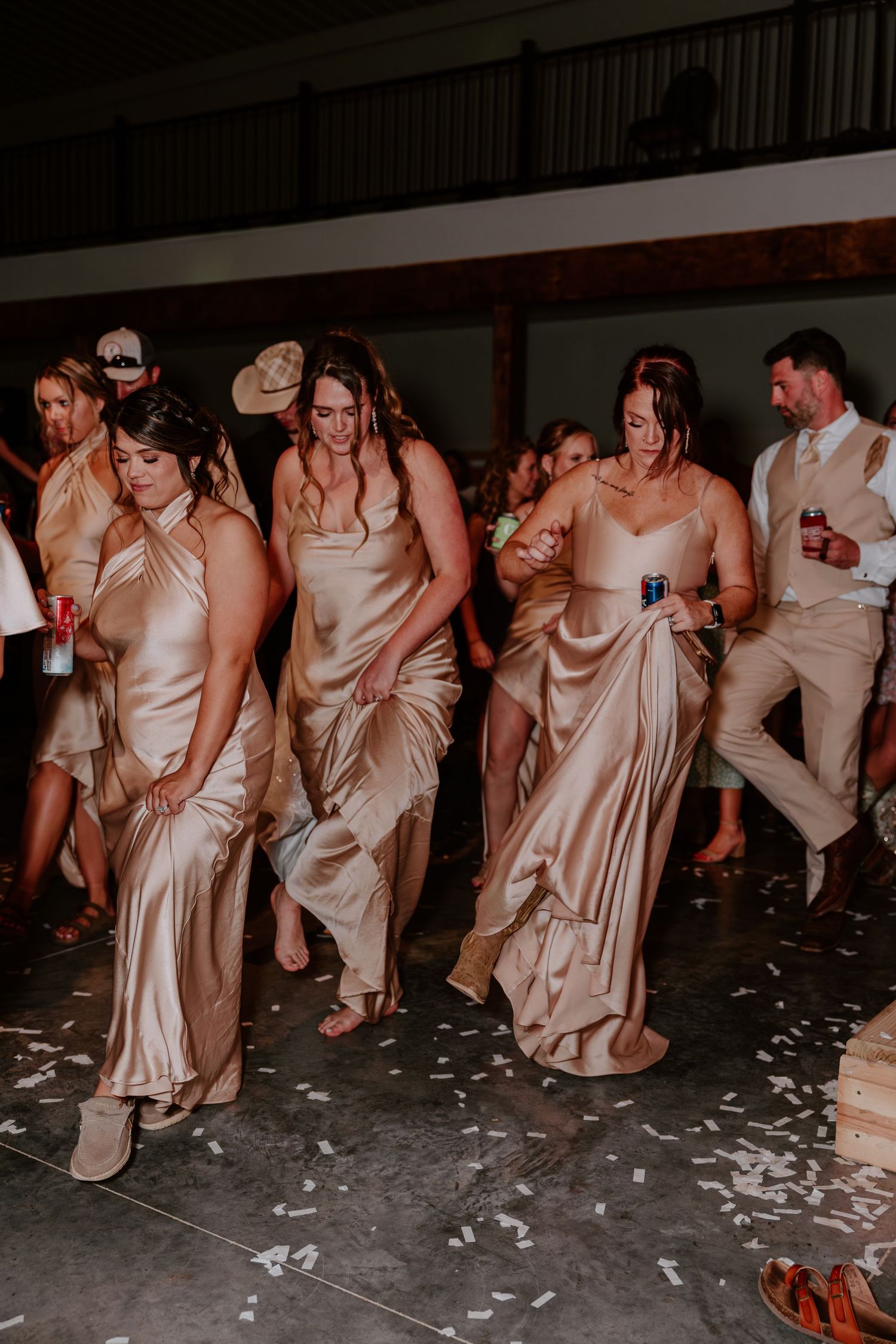 Bridesmaids in gold satin dresses dancing at a wedding reception on a confetti-covered floor.