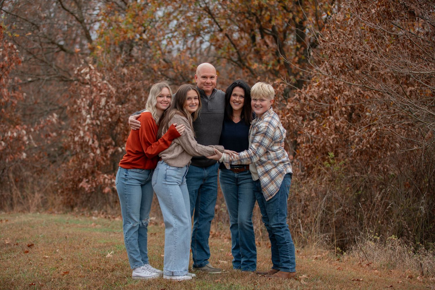 A family group poses together outdoors during autumn with fall foliage in the background.