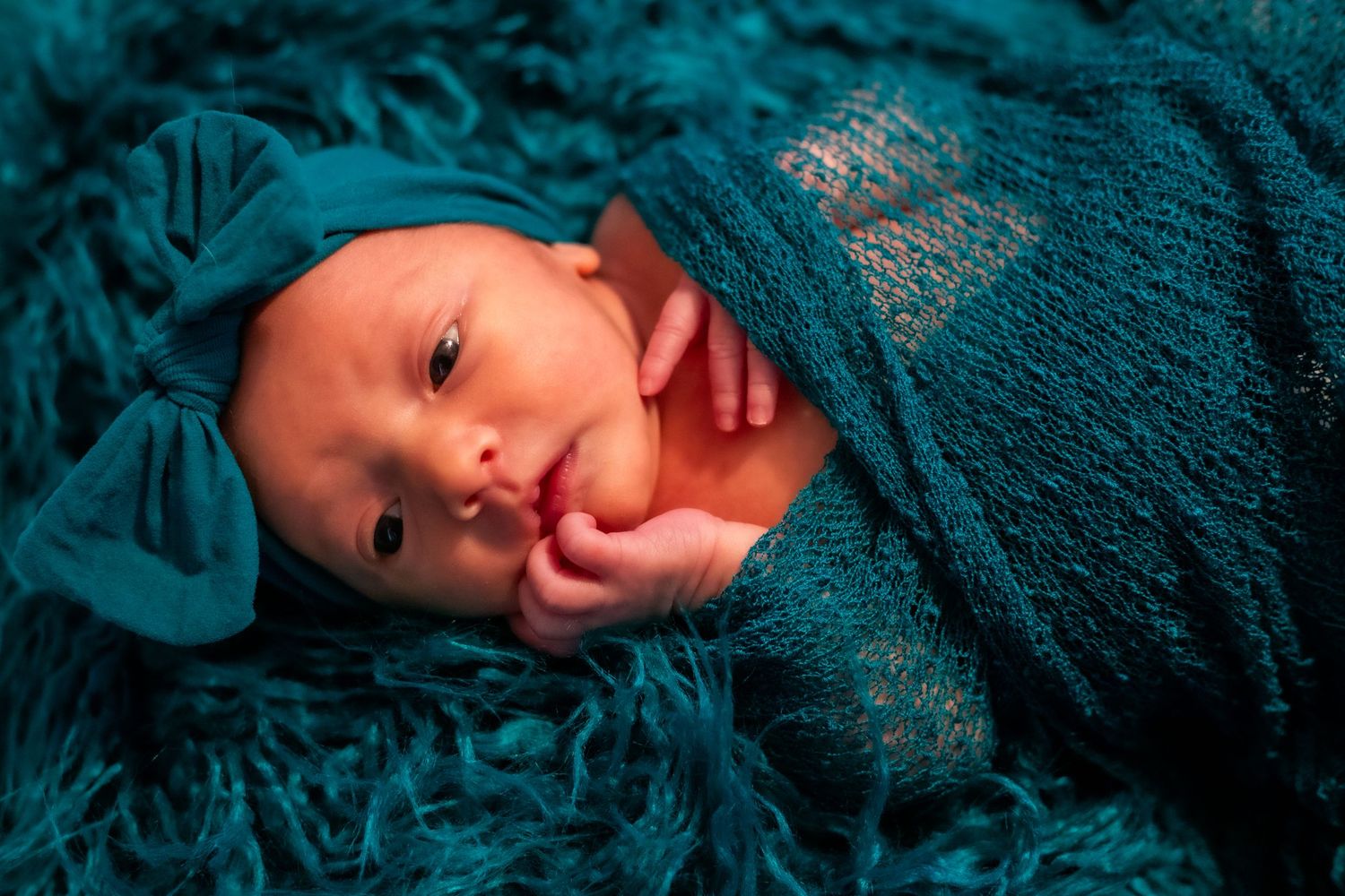 Newborn wrapped in teal blanket smiles against turquoise textured background.
