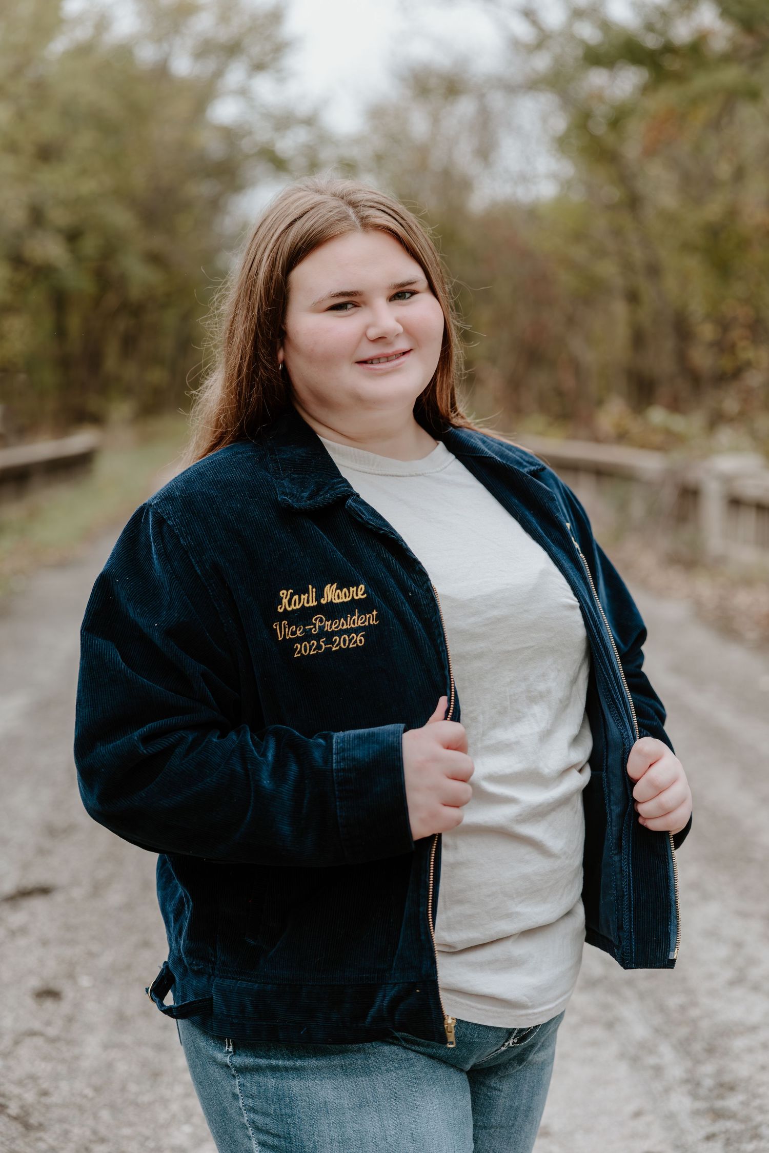 Person wearing a navy blue varsity jacket and light colored shirt stands on a gravel path in a natural outdoor setting.