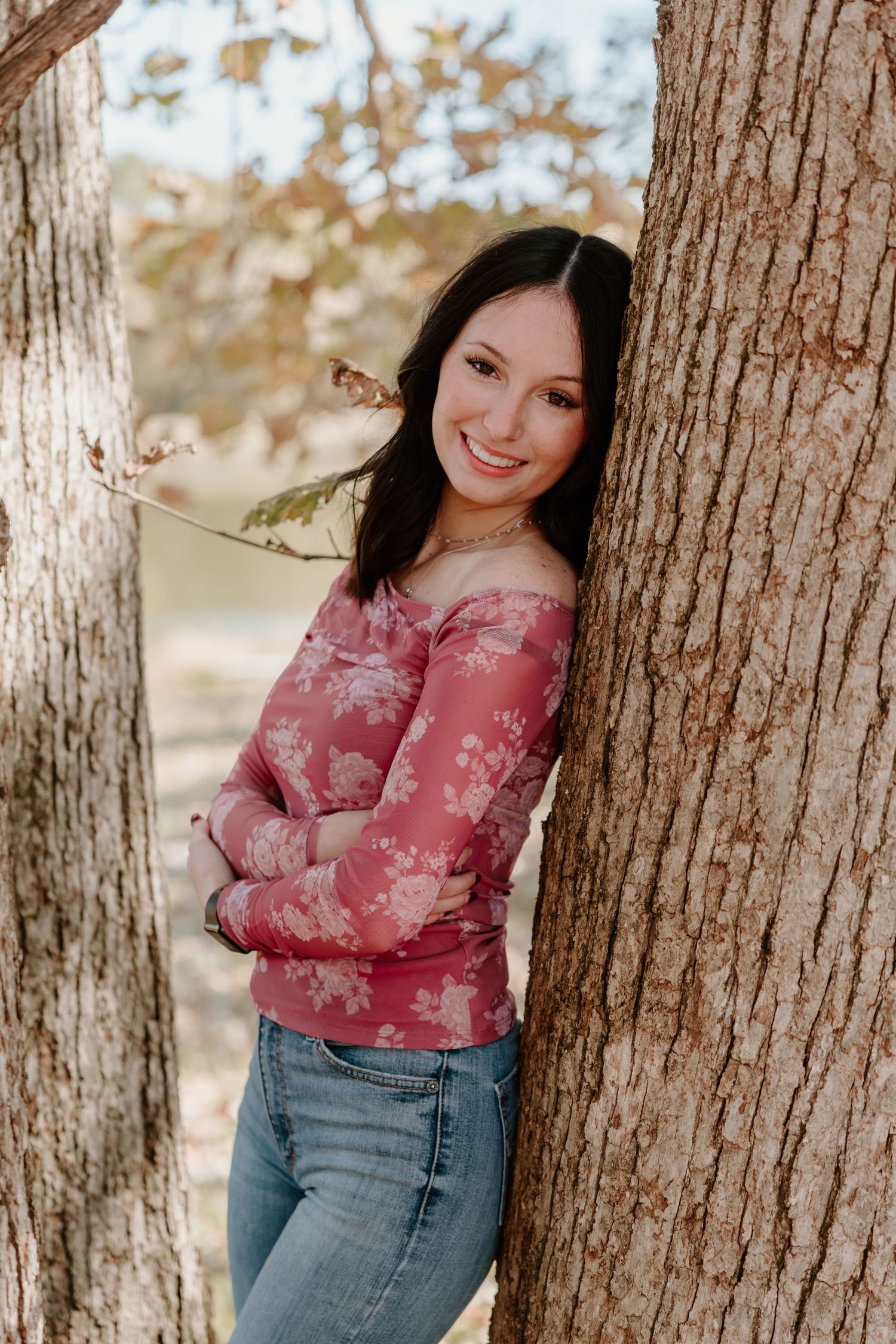 A person in a pink floral top and jeans poses casually against a tree trunk in an outdoor setting.