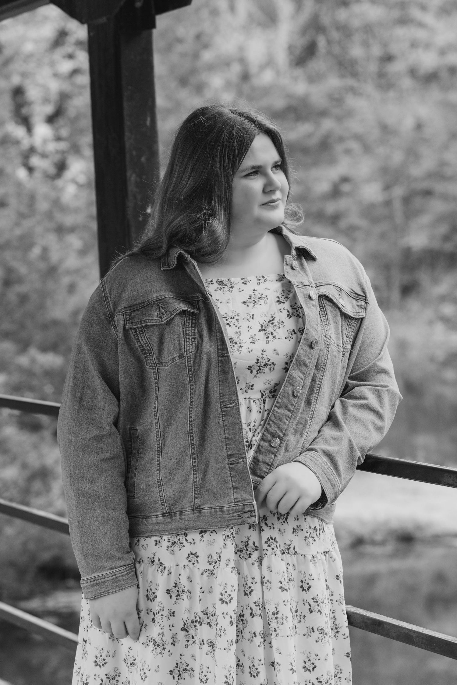 A person wearing a floral dress and denim jacket poses thoughtfully along a railing in a black and white photo series.