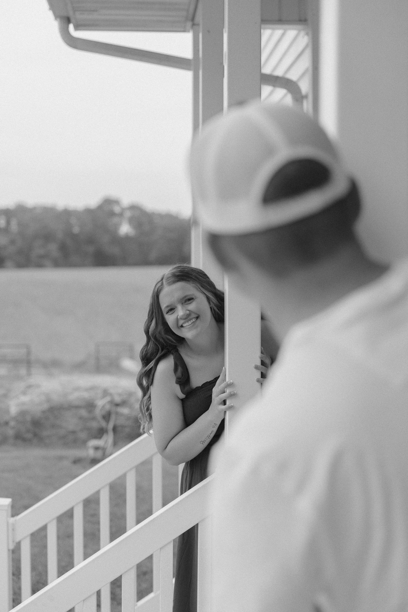 Black and white photo of a smiling woman peeking around a porch column at a man in a cap.