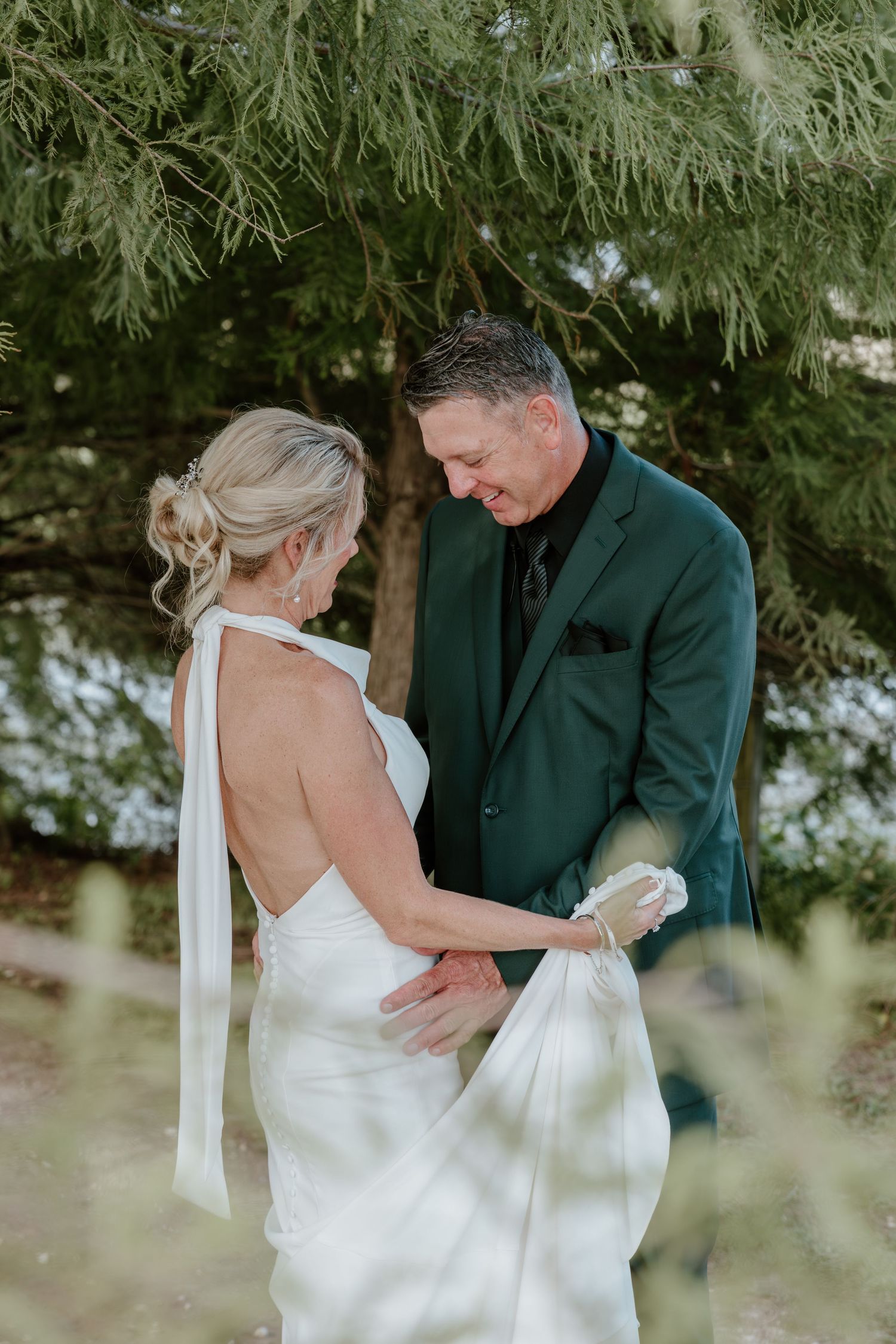 Groom in green suit holds bride's white gown during outdoor first look ceremony under pine trees.