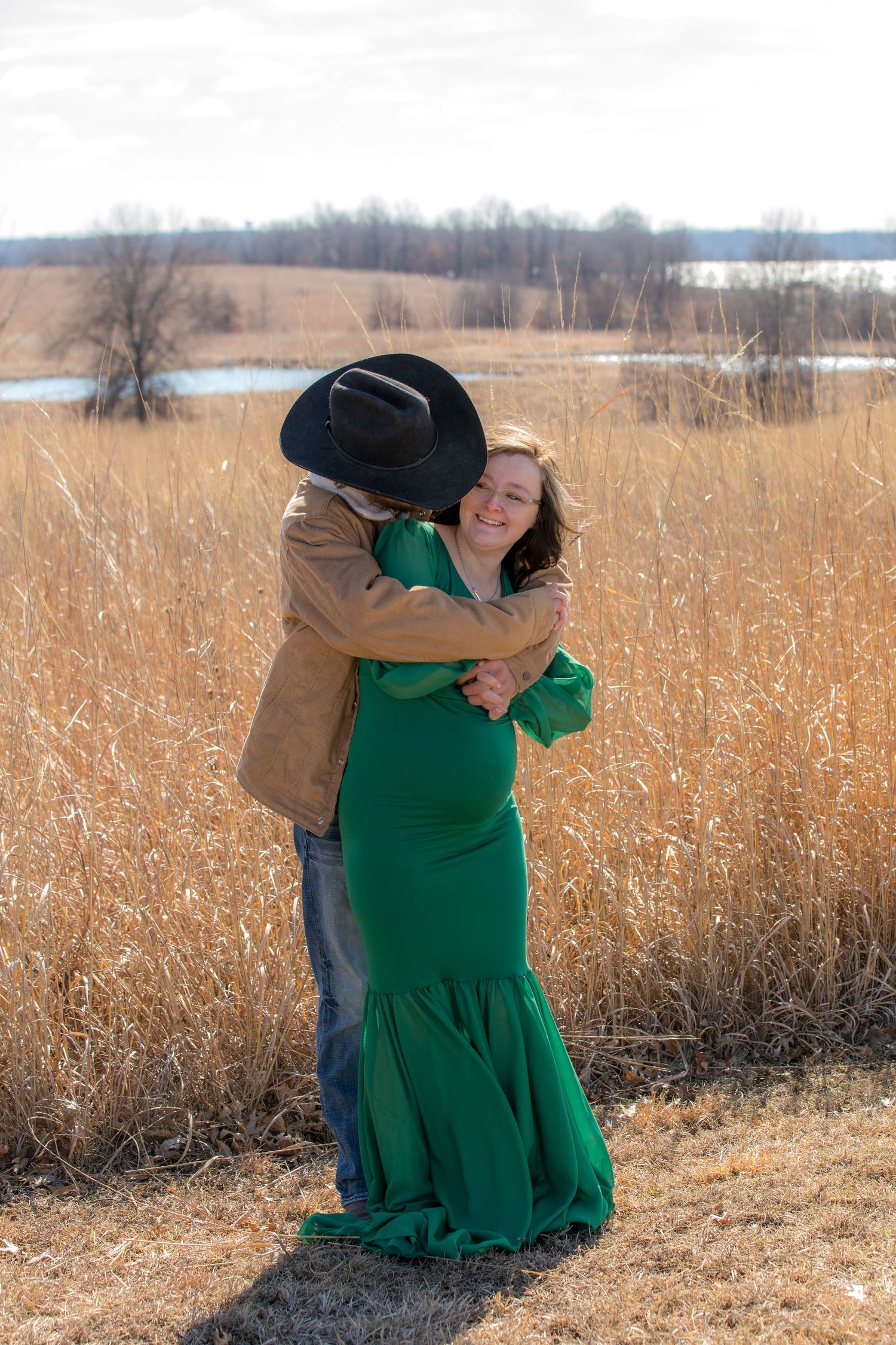 A couple embraces in a field of golden grass with water and trees in the background.