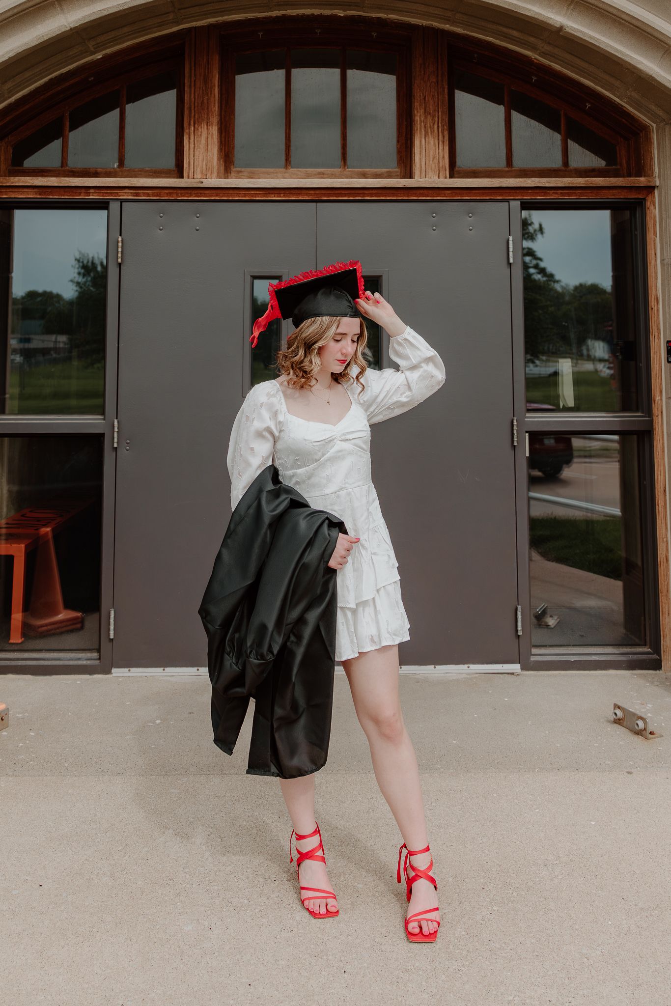 Young woman in white dress and red heels holds graduation gown, wearing black cap with red tassel outdoors.