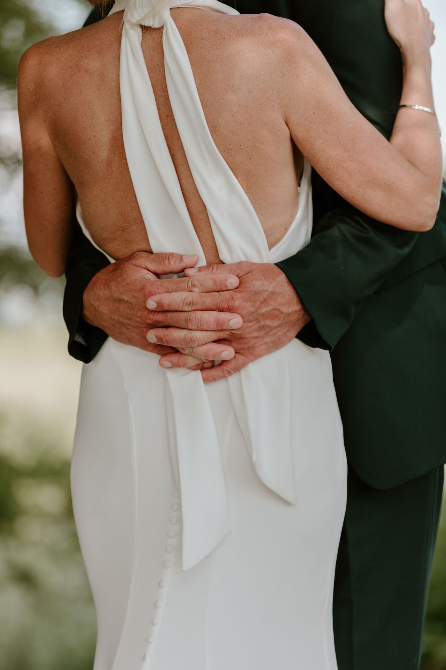 Bride in elegant backless halter wedding gown embraced by groom in dark suit at outdoor wedding ceremony.