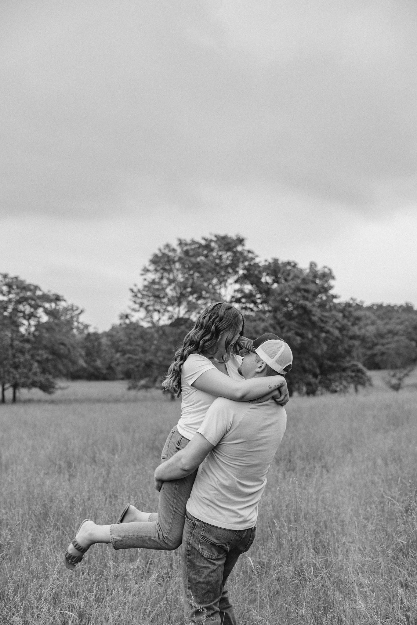Black and white photo of a couple embracing in a field, man lifting woman as they hug romantically outdoors.