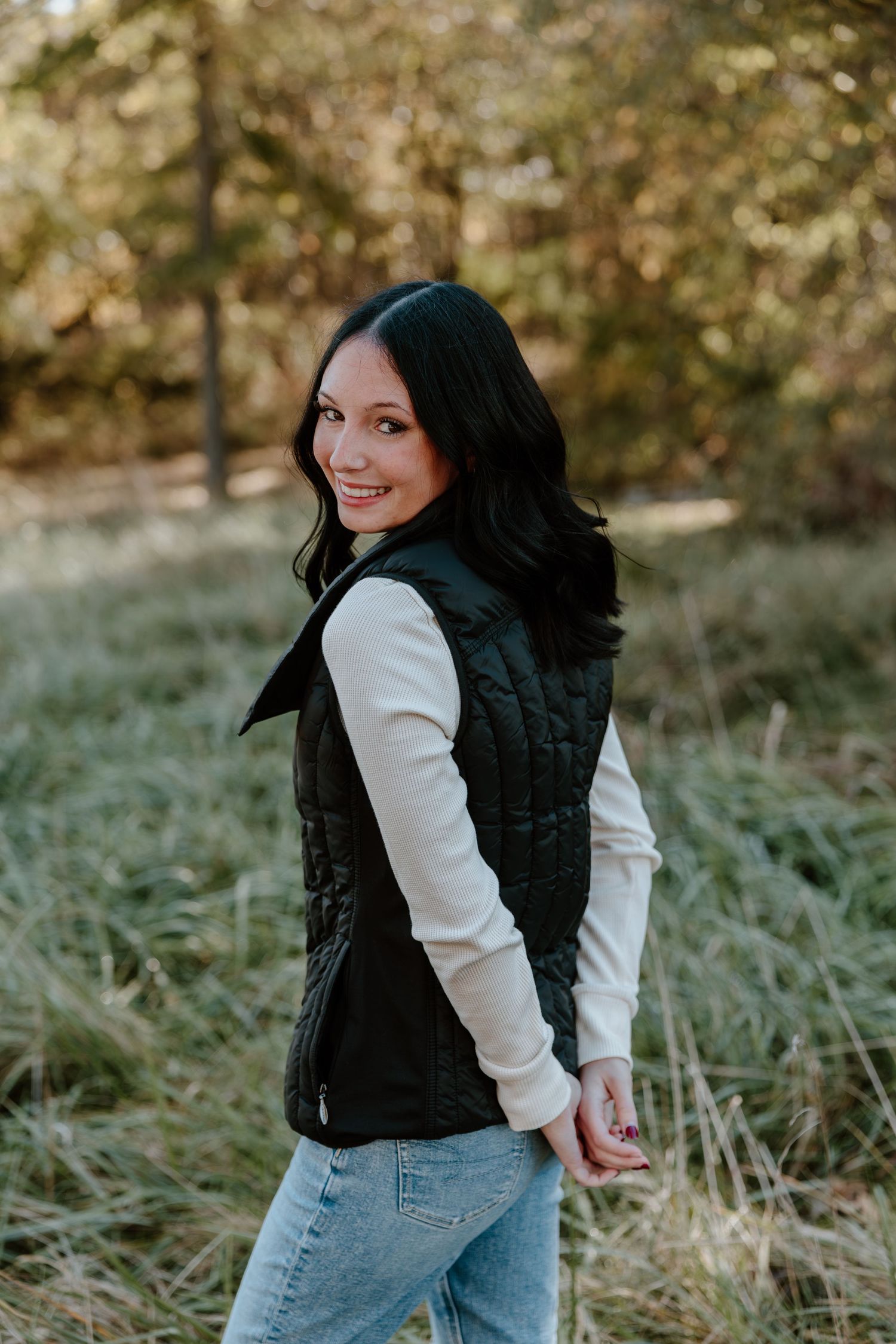 A woman in a black vest and white sweater poses playfully in a grassy outdoor setting during autumn.